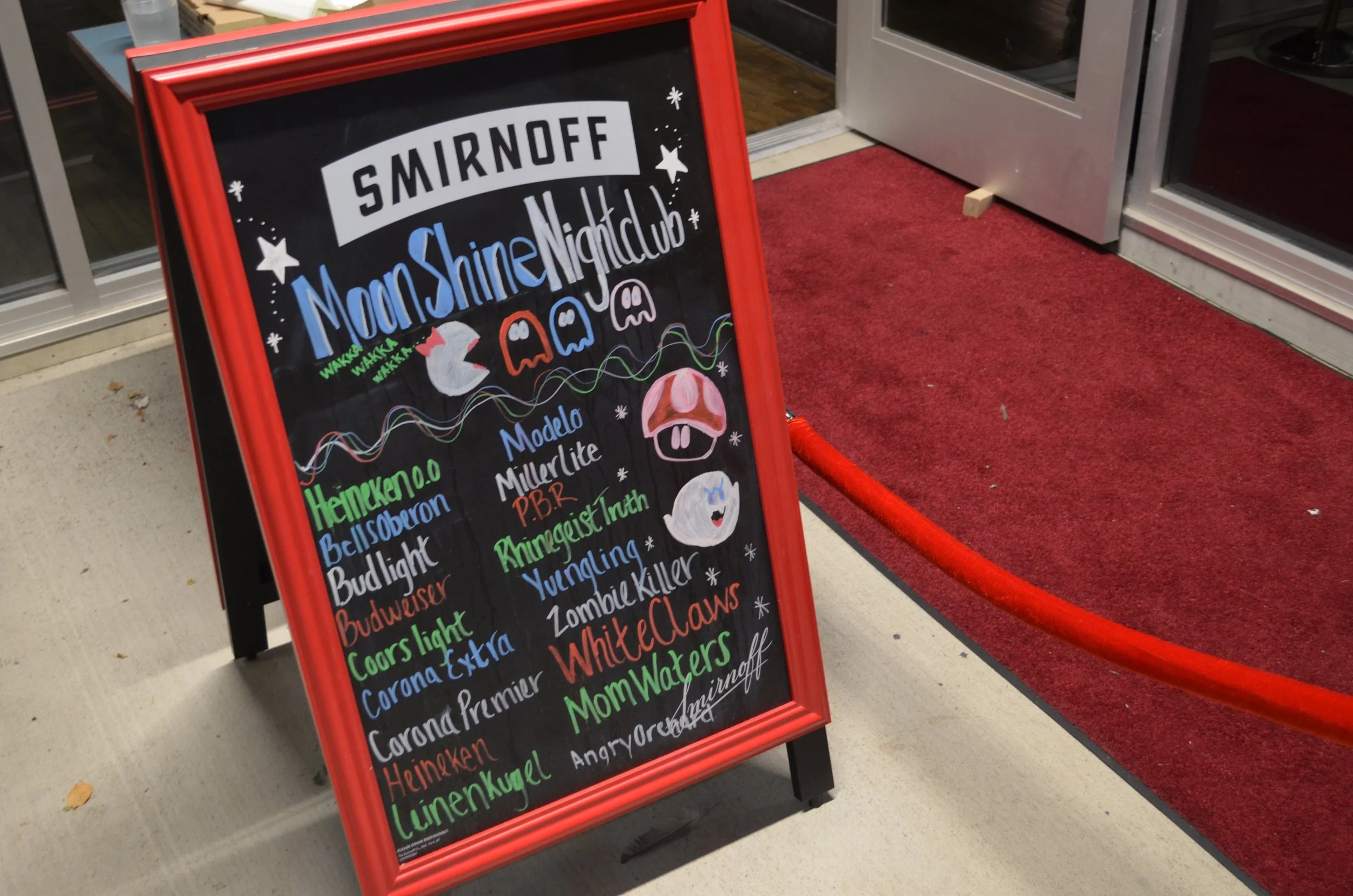 A blackboard sign with a red frame advertising a Smirnoff Moonshine Nightclub event, featuring themed drinks with colorful handwritten text and drawings of Pac-Man ghosts and characters.