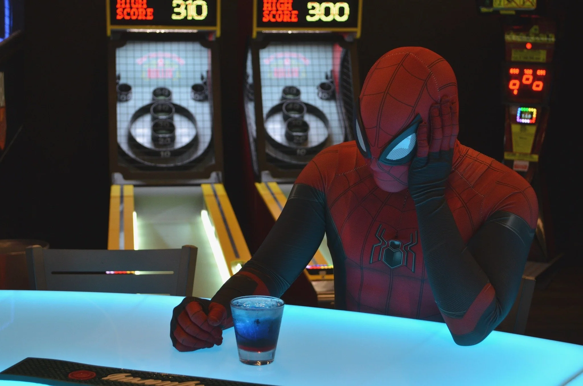 Person dressed as Spider-Man sitting at a bar with a drink in front of him, holding his head with his left hand, with arcade games in the background.