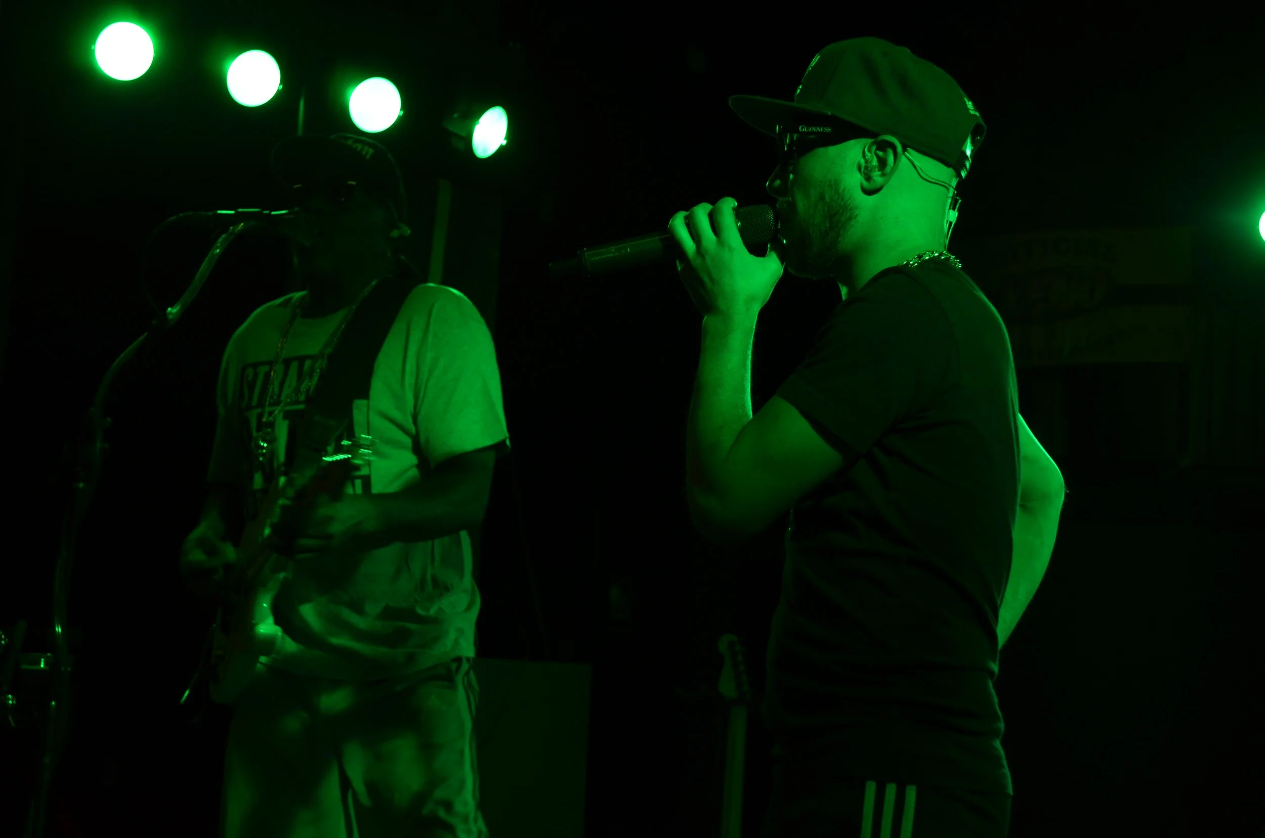 Two men performing on stage, with green stage lighting. One man is singing into a microphone, wearing a cap, sunglasses, and a T-shirt. The other man appears to be playing a guitar, wearing a cap and a T-shirt. The scene is dark with focused lighting