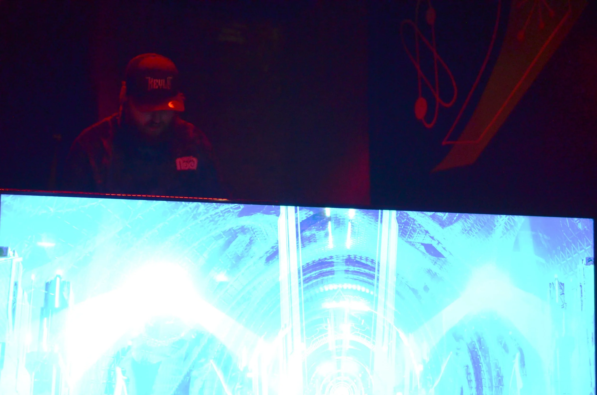 Person with a beard wearing a cap and dark clothing, looking down, illuminated by a bright blue and white light source, with abstract art or neon designs in the background.