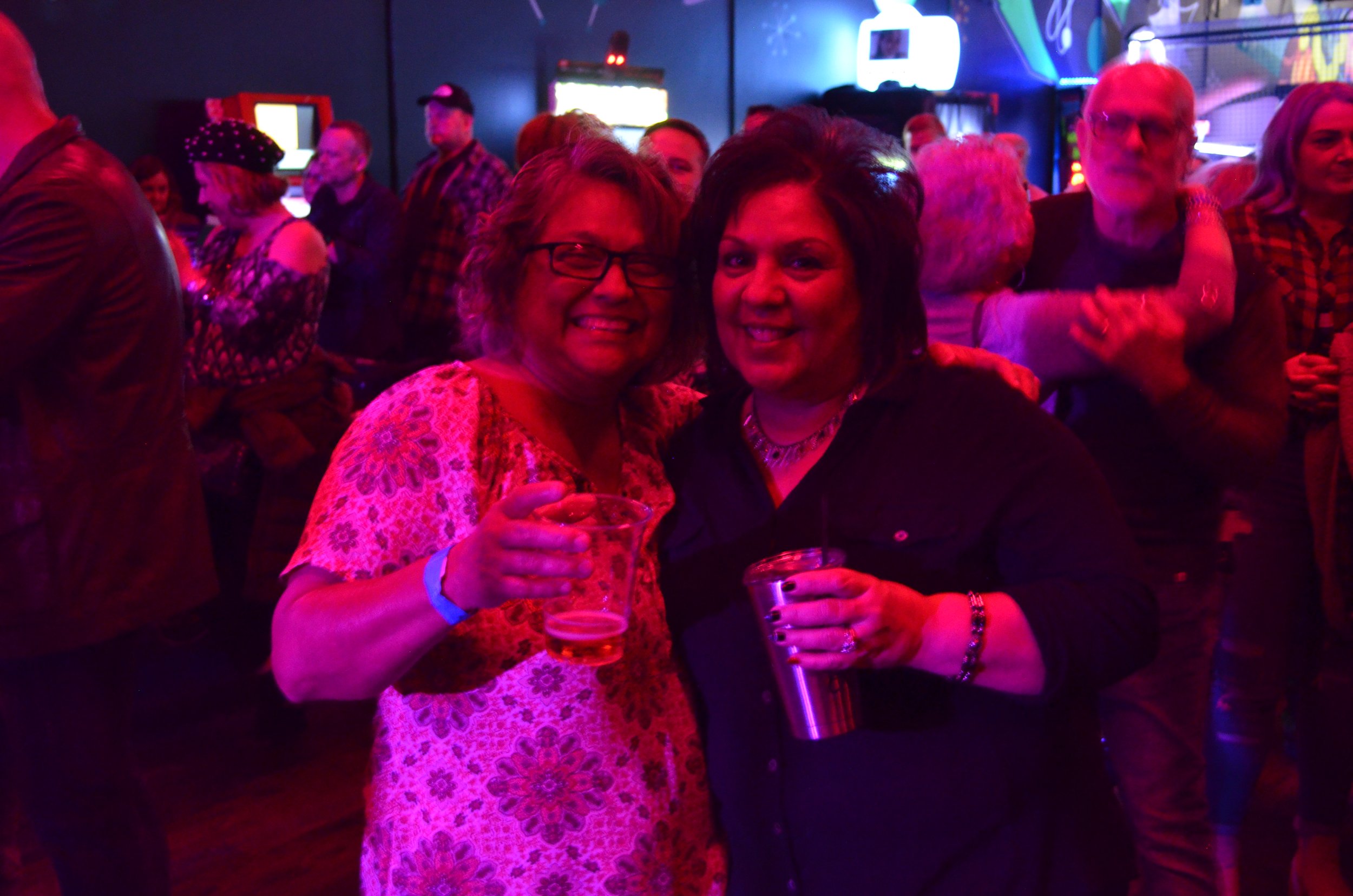 Two women smiling and holding drinks at a lively indoor party with colorful neon lights, surrounded by other people dancing and socializing.