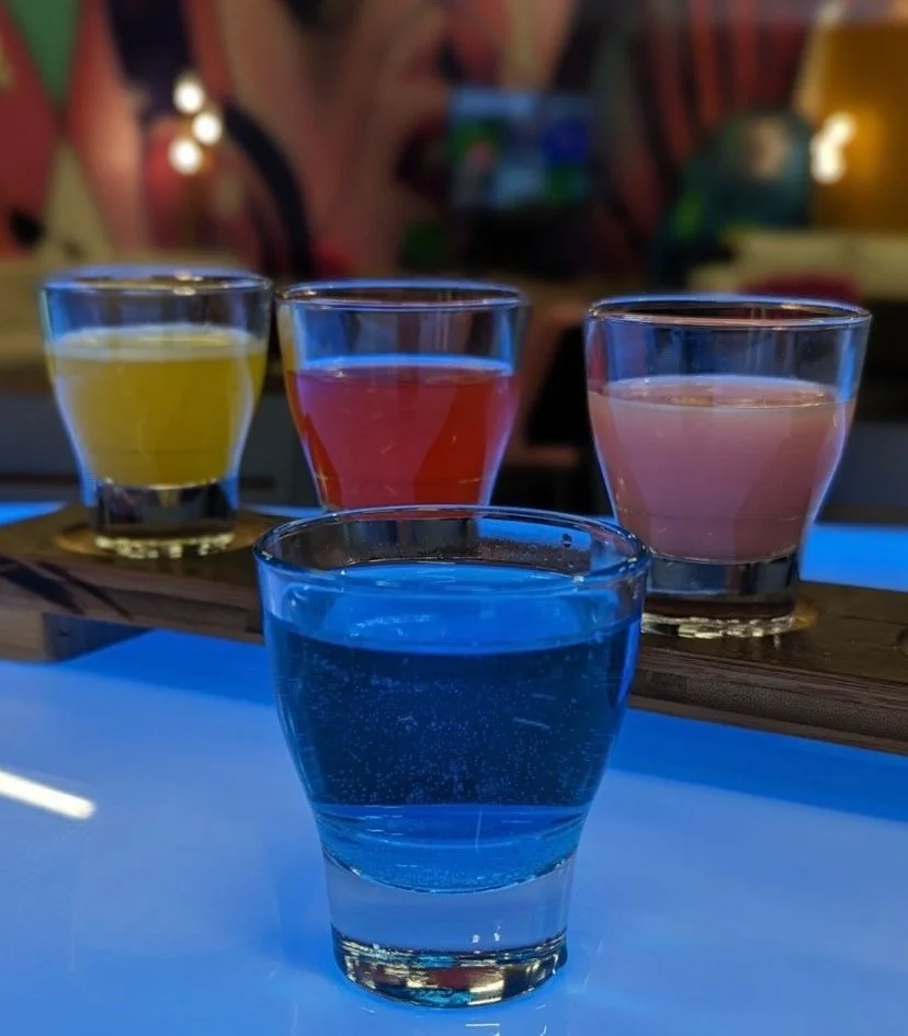 Four shot glasses filled with different colored liquids—yellow, red, pink, and blue—placed on a wooden surface with a blurred colorful background.