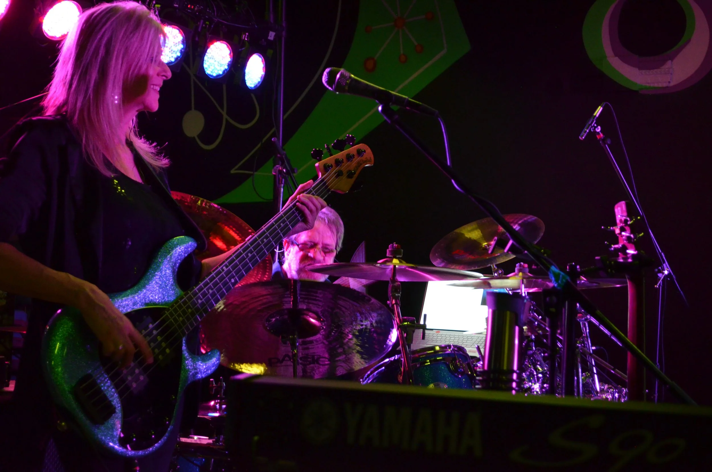 Female guitarist with a glittery green bass guitar and male drummer playing a drum set on stage with colored lights.