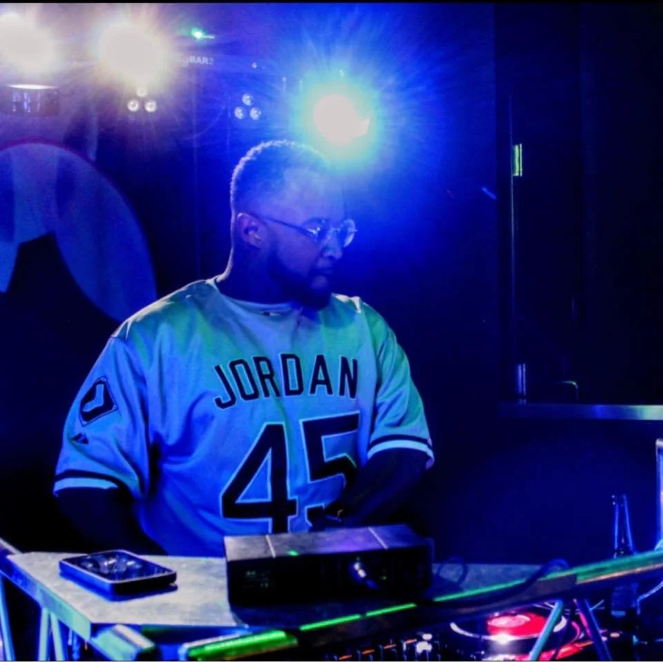 A man with glasses and a beard wearing a white Jordan football jersey with the number 45 and the name Jordan, standing at a DJ setup with colorful blue and green lighting.