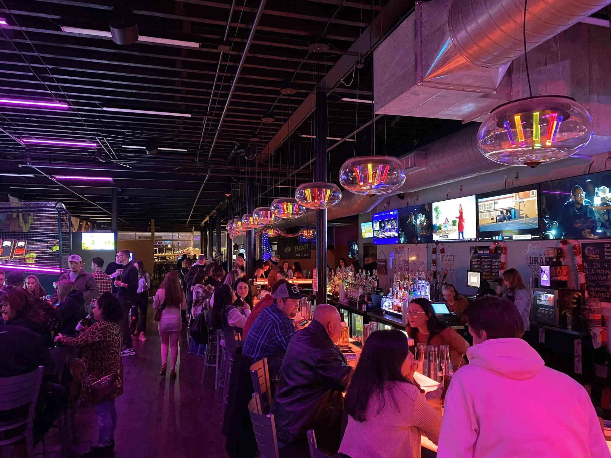 A lively bar with people socializing and ordering drinks, illuminated by colorful neon and LED lighting.