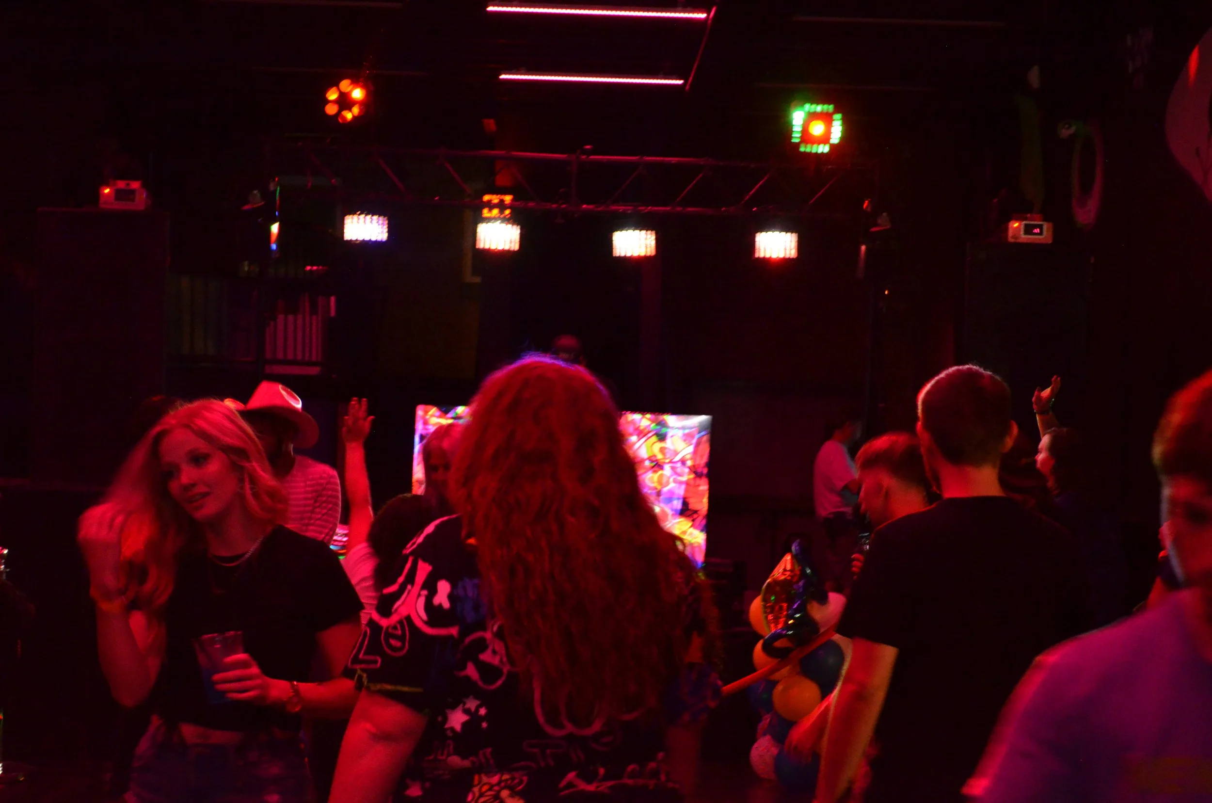 People dancing and socializing in a nightclub with colorful lighting and a DJ in the background.