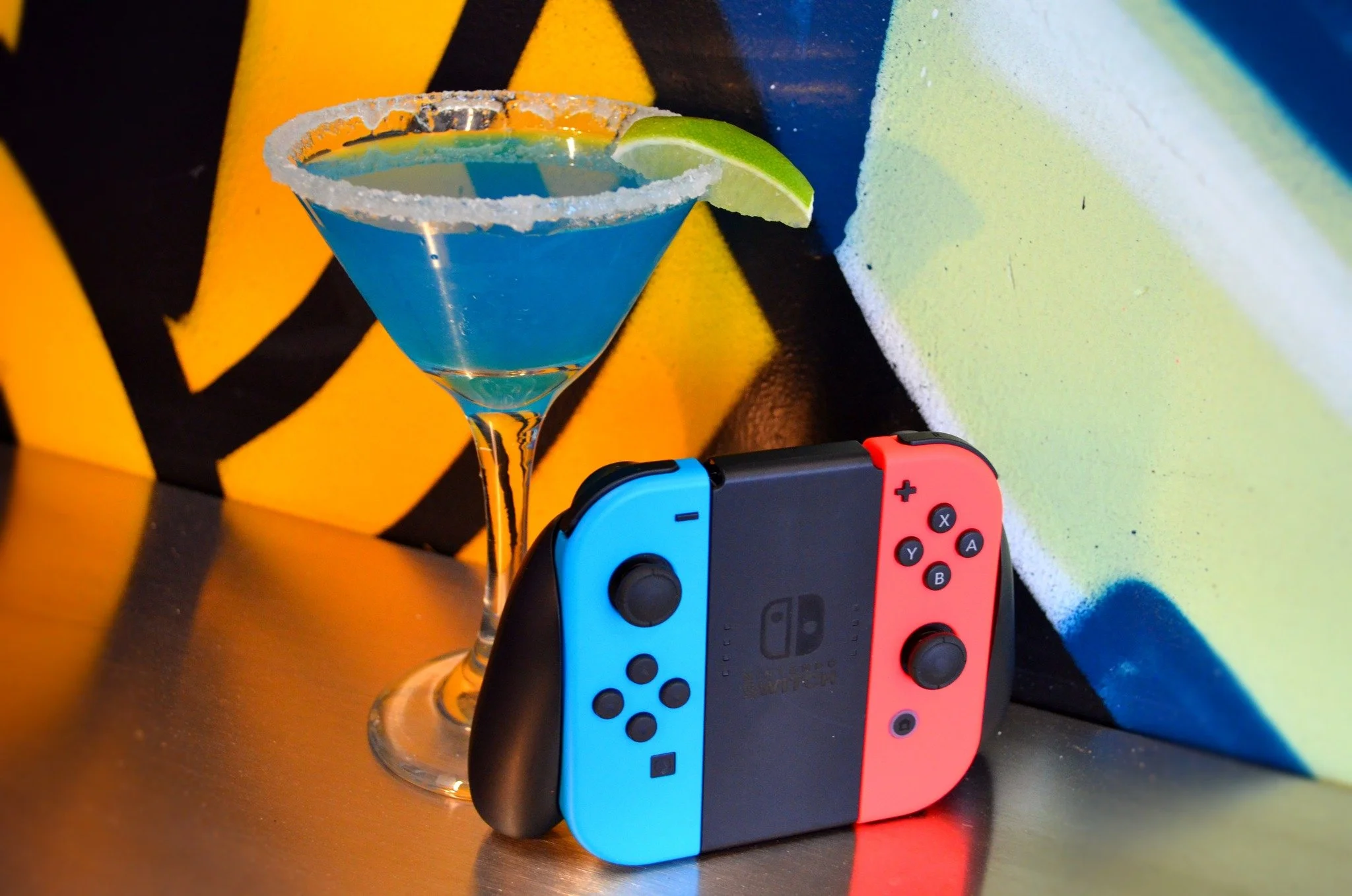 A blue cocktail in a martini glass with a lime wedge and salted rim, and a Nintendo Switch console with blue and red controllers on a metallic surface, against a colorful graffiti wall background.