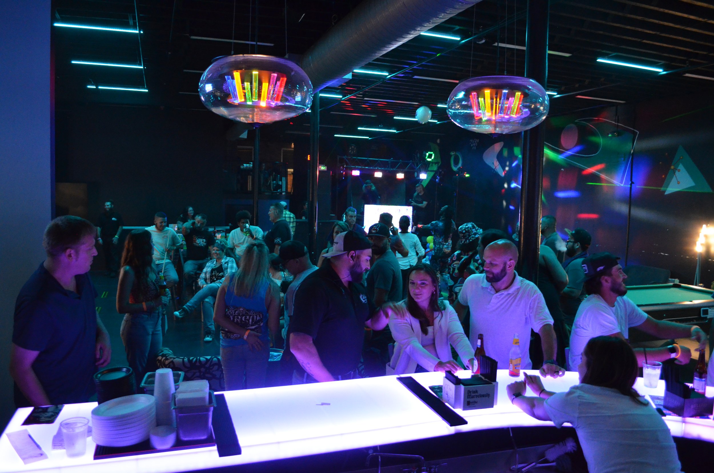 People dancing and socializing at a nightclub with neon lights and colorful laser effects.