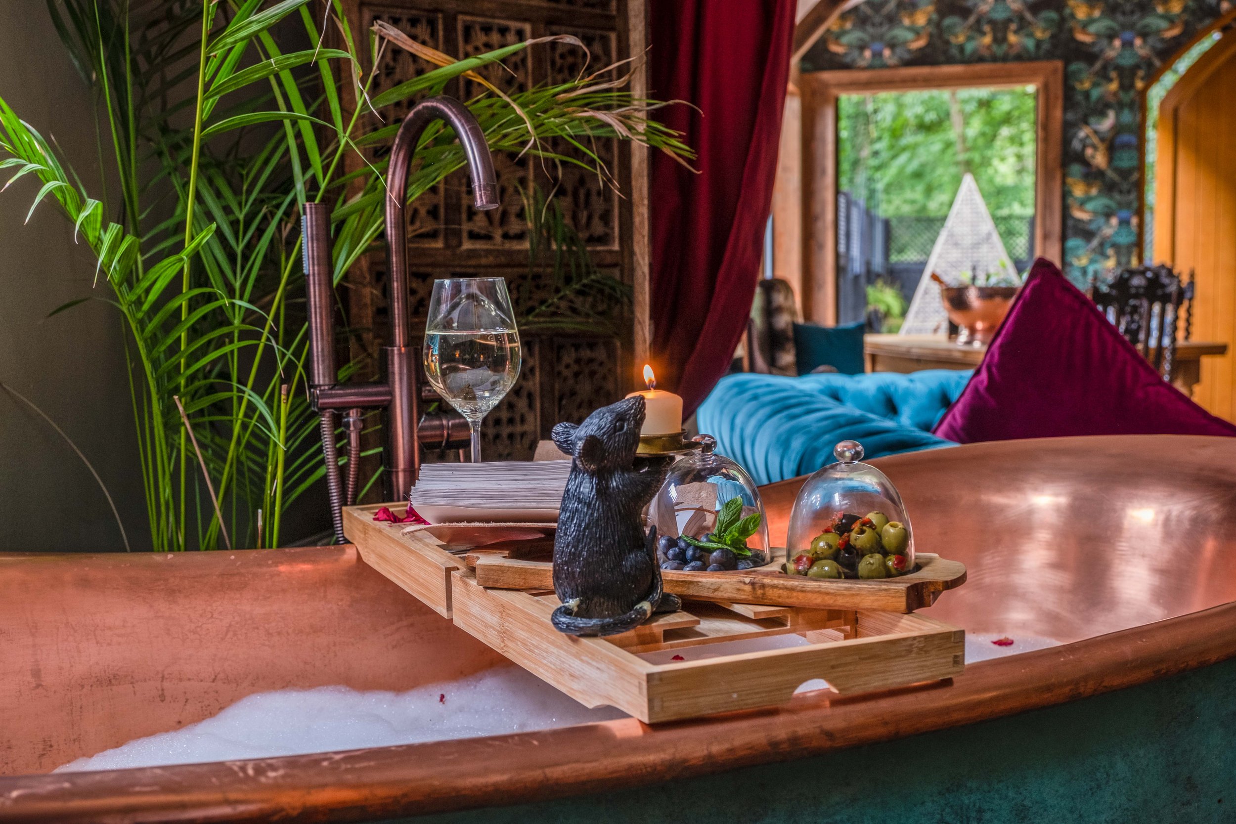 Luxurious copper bathtub with bubble bath, wooden tray holding wine glass, candle, and covered dishes, green indoor plants, elegant interior decor with blue sofa and purple pillows in background.