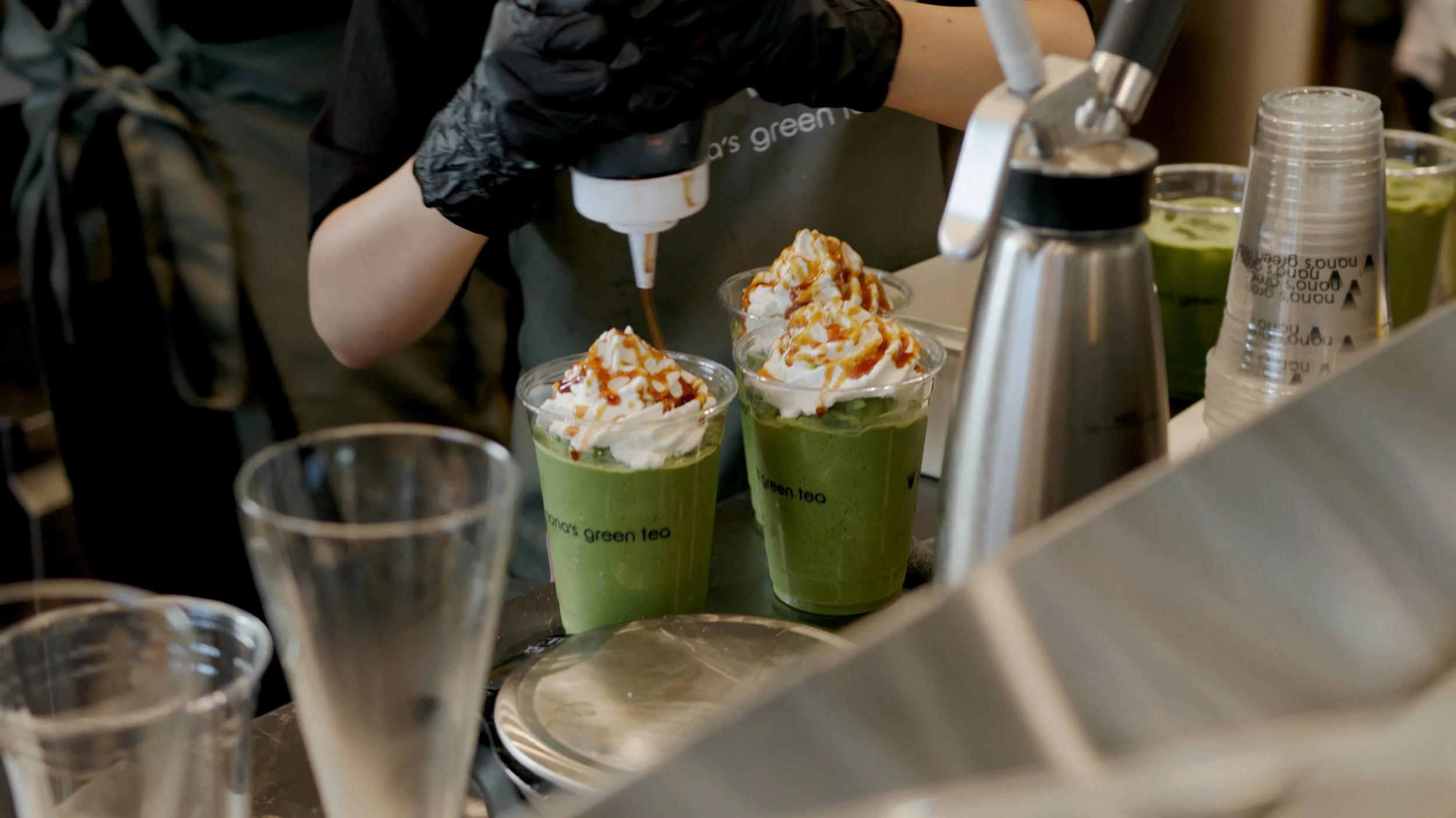 A worker preparing matcha green tea drinks topped with whipped cream and caramel drizzle in clear plastic cups at a cafe or coffee shop.
