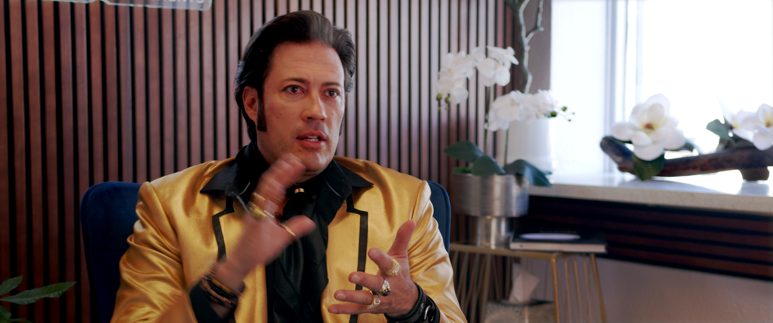 A man with styled dark hair and sideburns, wearing a shiny gold jacket over a black shirt, gesturing with his hands while sitting on a blue chair in a room with wooden wall paneling and white orchids on a windowsill.