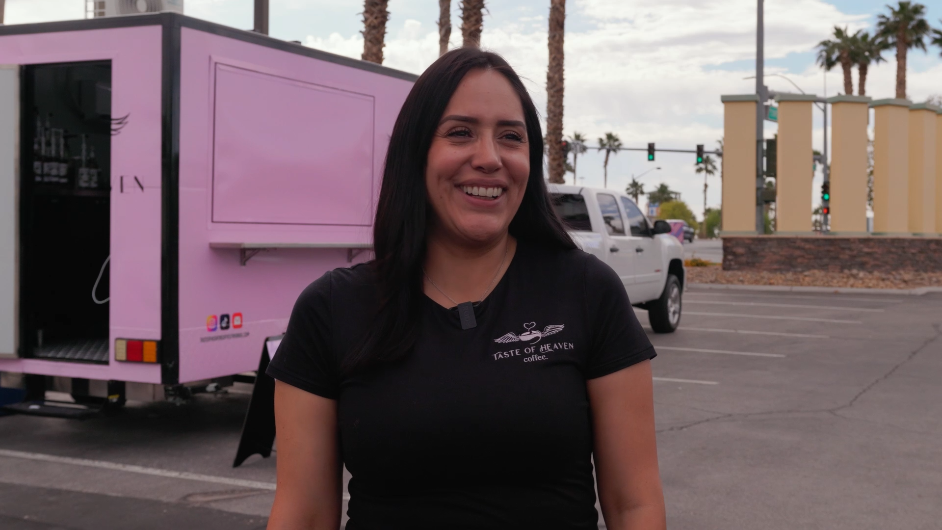 A woman with long dark hair smiling in a parking lot, wearing a black T-shirt that says 'Taste of Heaven Coffee,' with a pink food truck and palm trees in the background.
