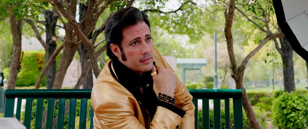 A man with styled black hair and sideburns sitting on a park bench, wearing a shiny gold jacket, black shirt, and bracelets, looking contemplative with trees and greenery in the background.