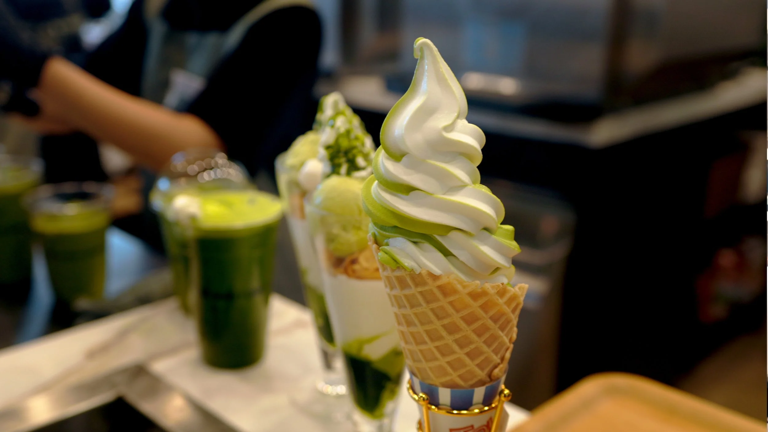 Soft serve ice cream cone with green and white swirl in focus, with other ice cream desserts and drinks with green toppings blurred in the background.