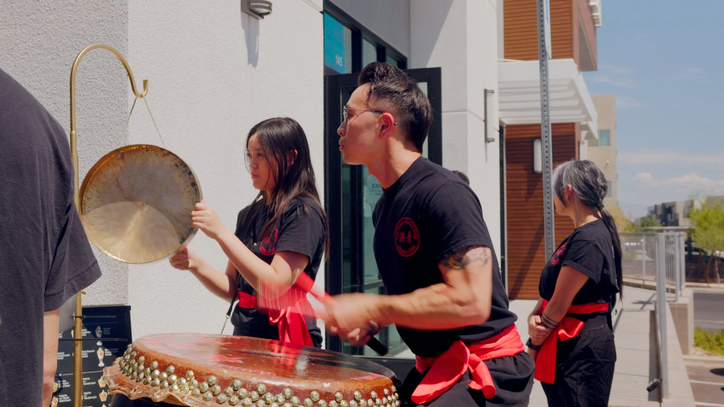 People performing Taiko drumming outdoors, wearing black outfits with red sashes, and playing large drums.