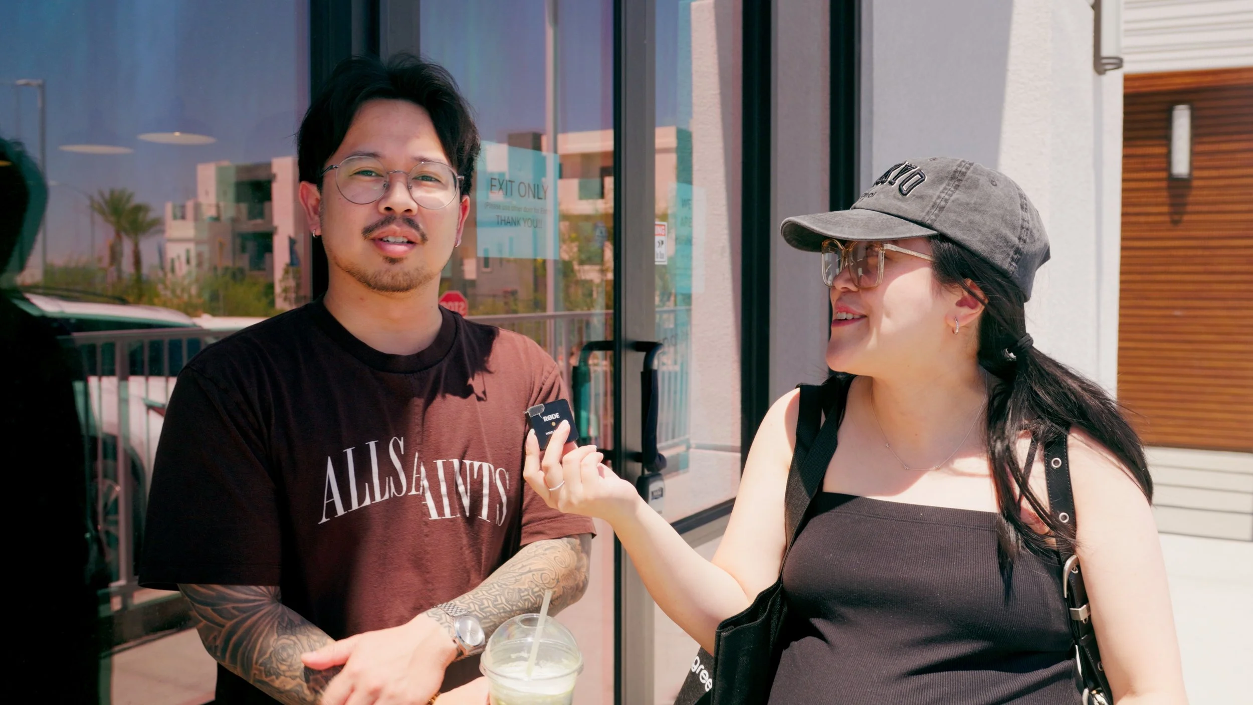 A man with glasses and tattoos is being interviewed by a woman holding a microphone outside a modern building on a sunny day.
