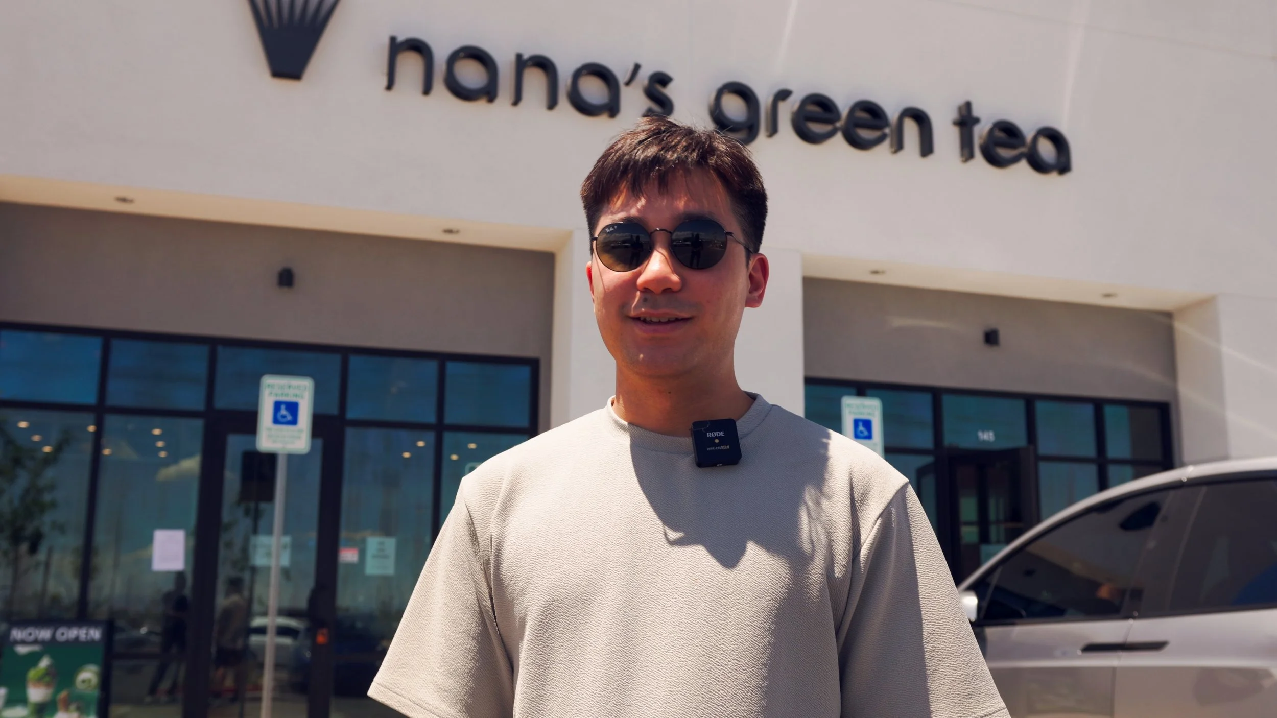 A young man wearing sunglasses and a beige t-shirt stands outside Nana's Green Tea store, smiling at the camera. There are handicap parking signs and a car in the background.
