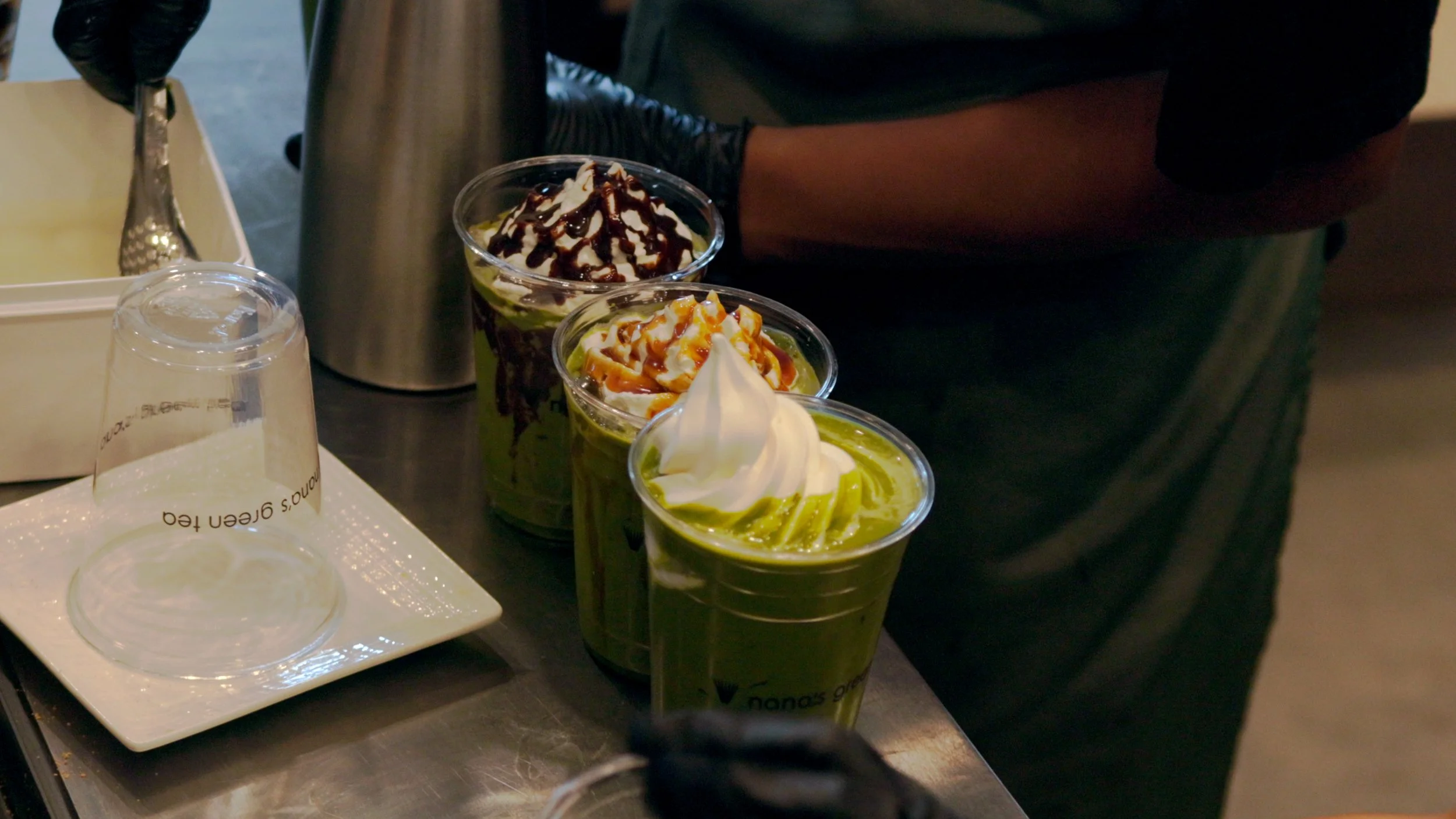 Three cups of matcha green tea lattes with whipped cream and caramel, topped with different sauces, on a countertop with a person in a black shirt and gloves in the background.