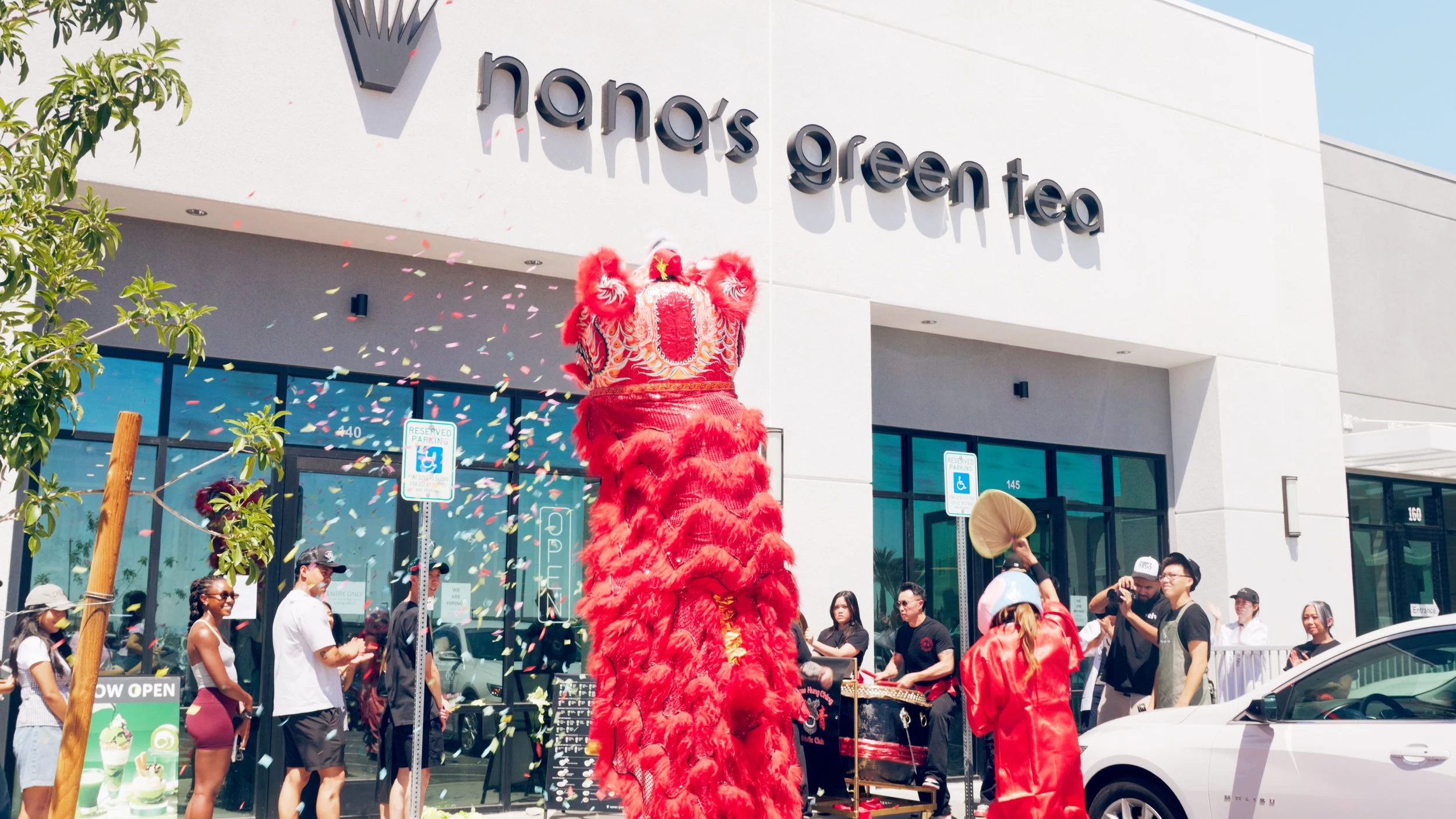 Traditional Chinese lion dance performance during grand opening event at Hanabrea Greentea storefront, with people watching and confetti falling from the sky.