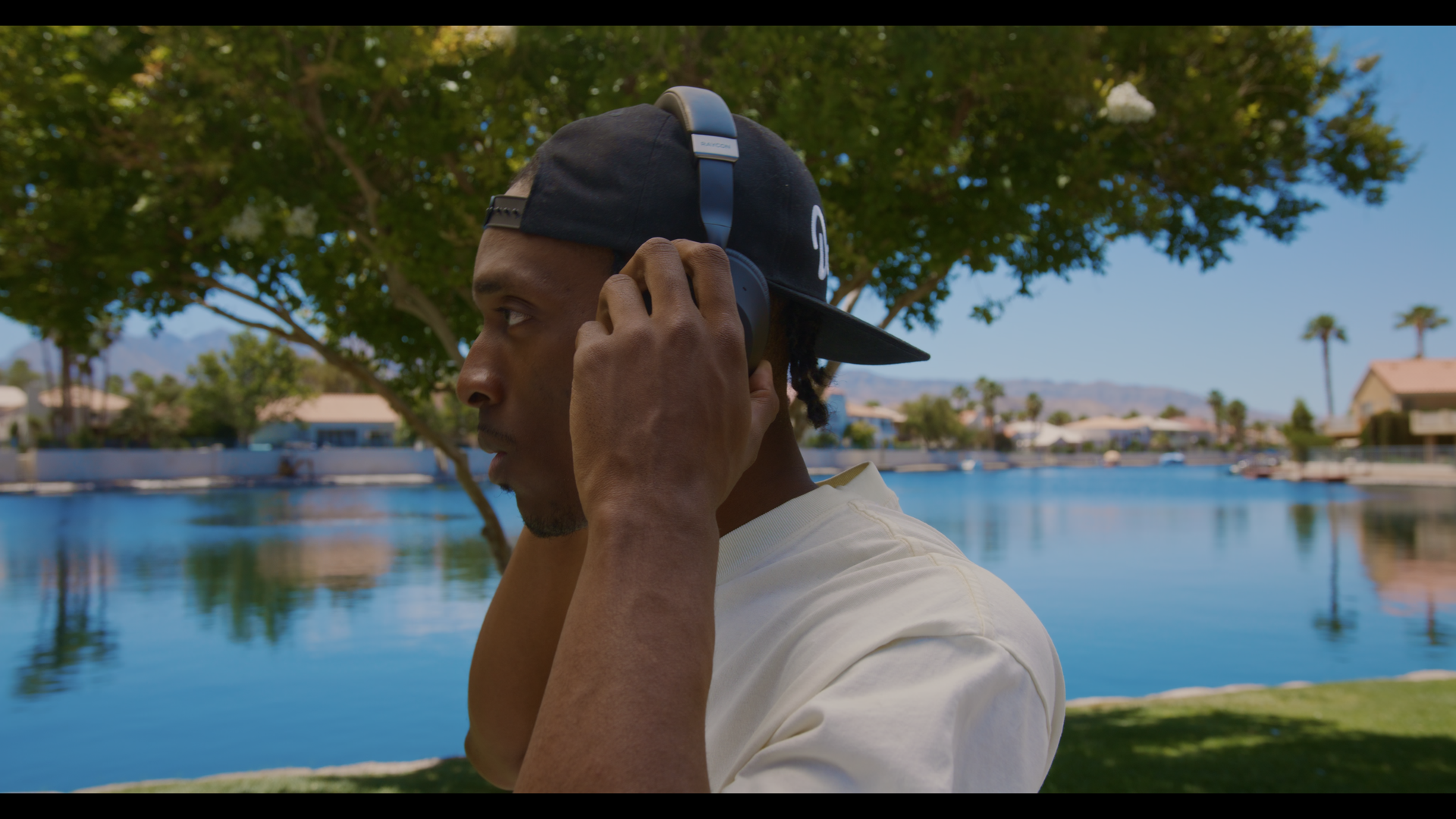 A young man wearing a black cap and white t-shirt is adjusting blue headphones on his head outdoors by a lake with trees, houses, and mountains in the background.