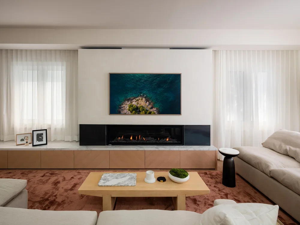 Modern living room with a fireplace, abstract artwork on the wall, white curtains, beige sofa, black accent table, and wooden coffee table with decorative objects.