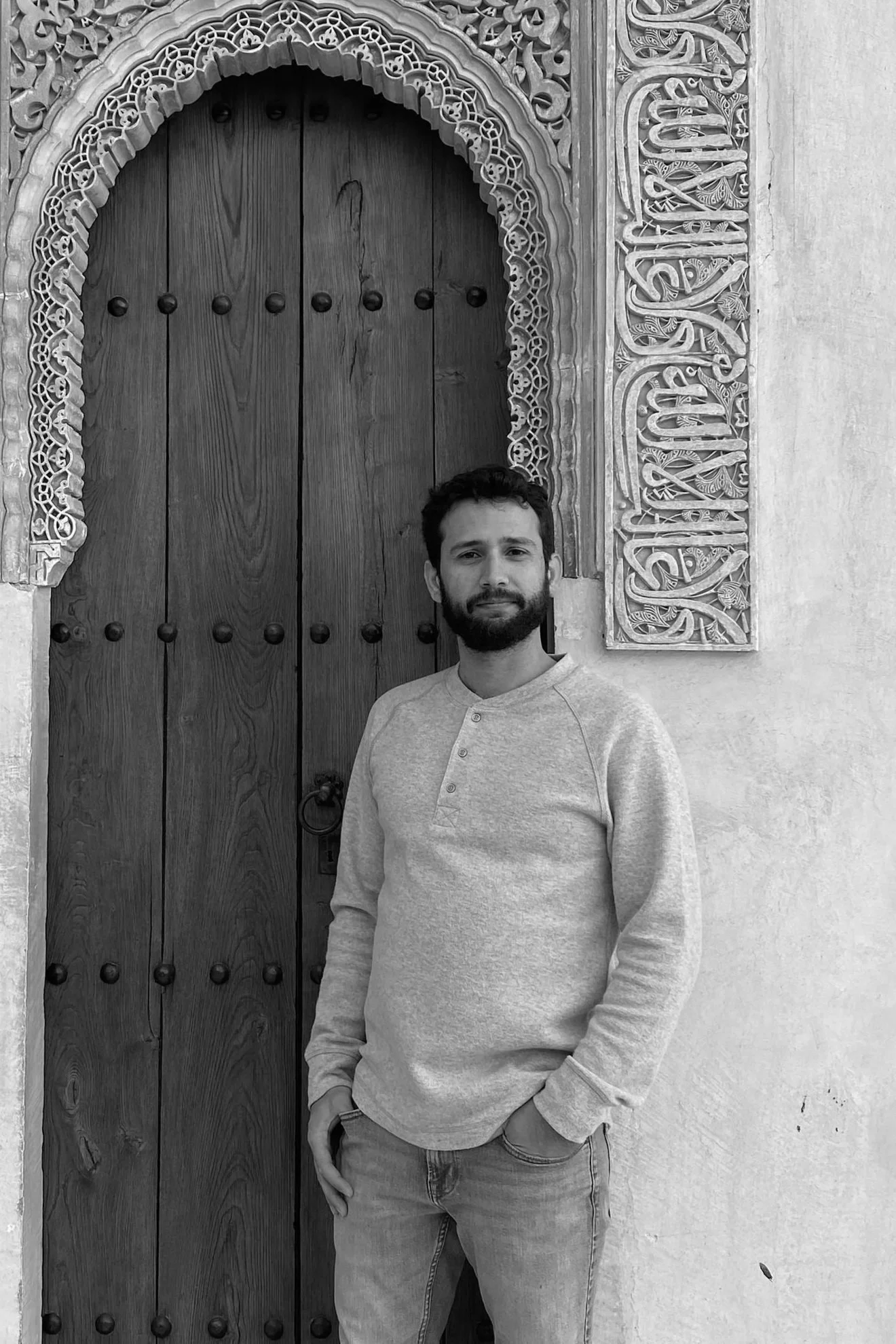 A man with a beard, wearing a light-colored sweatshirt, standing in front of a wooden door with decorative carved trim. The photo is black and white.