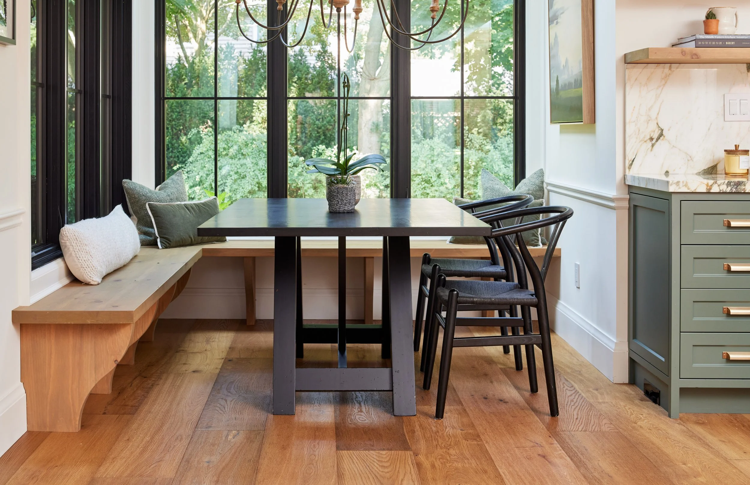 A cozy dining area with a wood bench on one side and a dark wooden table surrounded by black chairs. The space features large windows with black frames, allowing natural light and a view of lush green trees outside. There are decorative pillows on the bench and a potted plant on the table. The corner of a kitchen with mint green cabinets and a marble countertop is visible on the right, along with a wall-mounted television and shelves with books and small decor items.