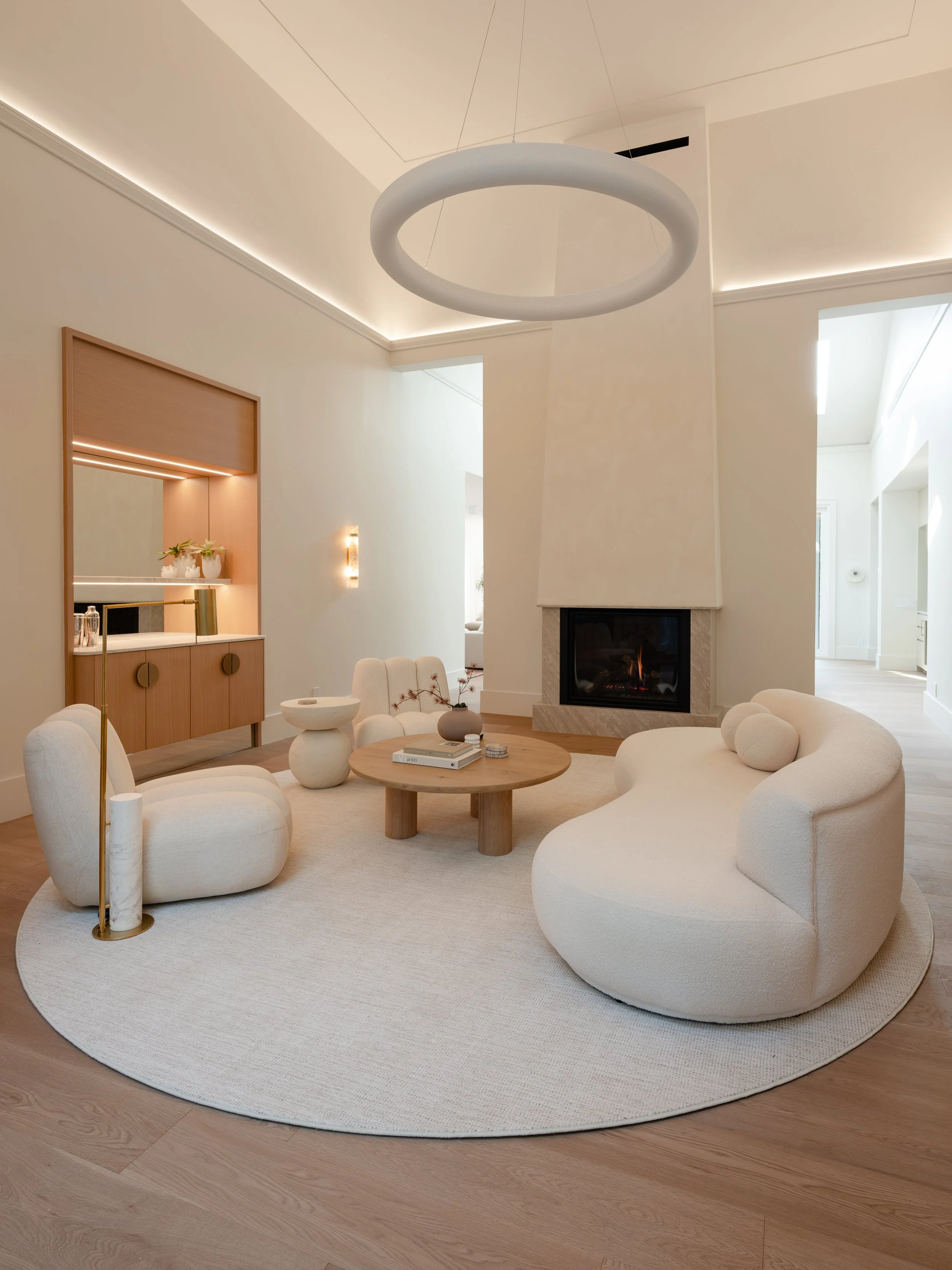 Modern living room with beige curved sofa and matching armchair, round wooden coffee table, white rug, fireplace, and large circular chandelier, with natural light coming through windows.