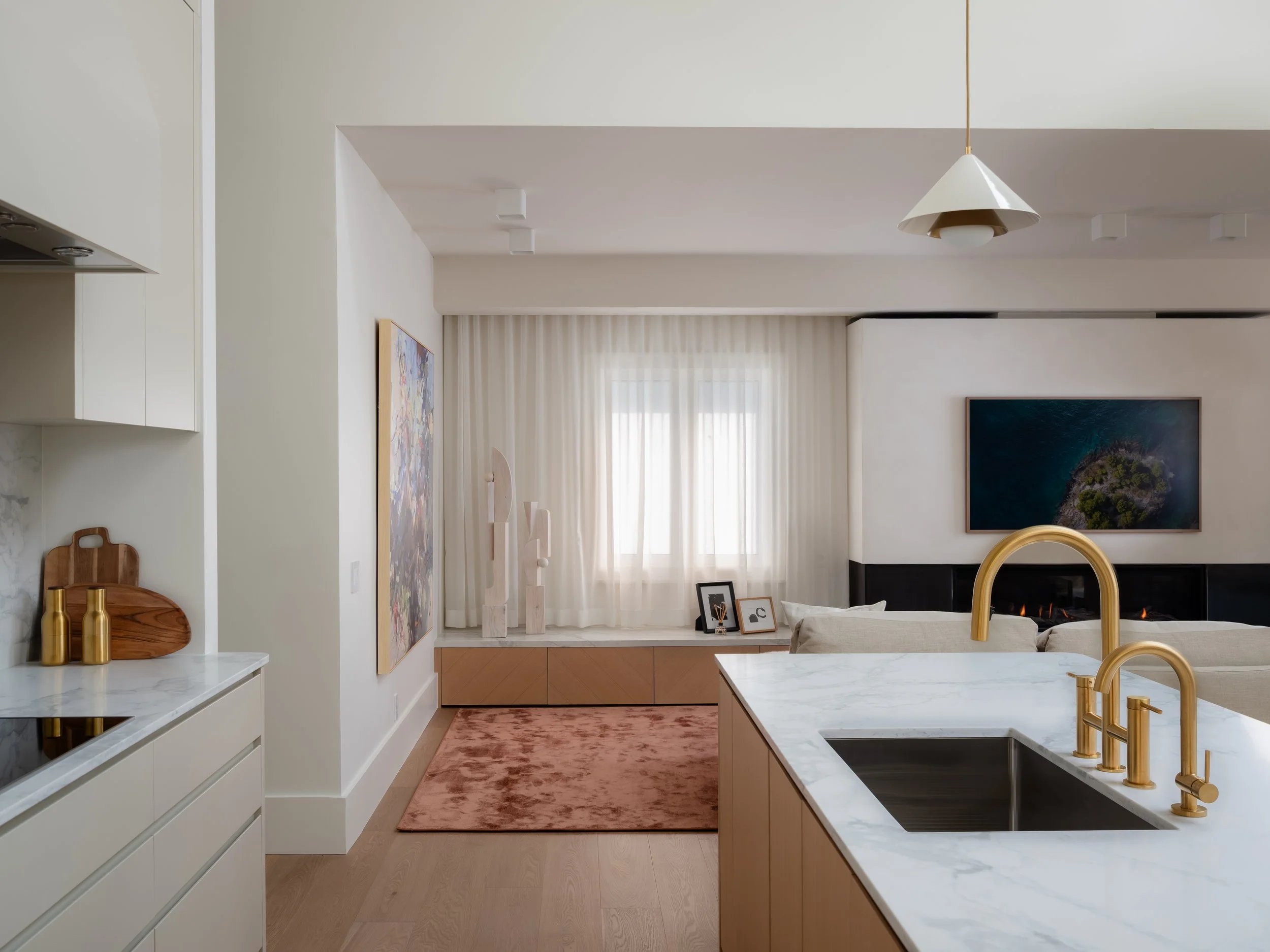 Modern kitchen with a white marble island, gold fixtures, and a view into a living area with artwork, a fireplace, and a window with curtains.
