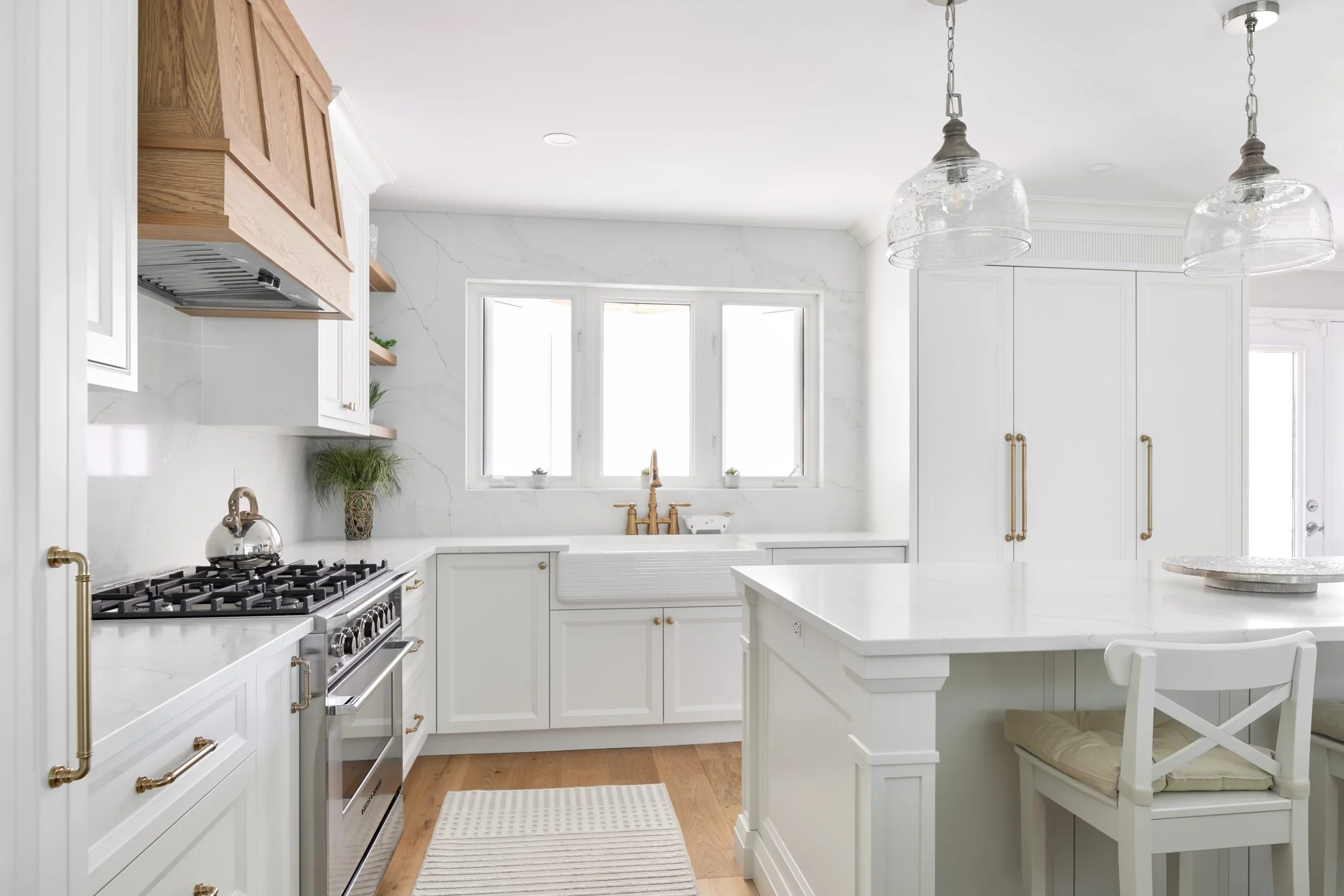 A bright, modern kitchen with white cabinetry, marble countertops, and a white farmhouse sink. There are large windows above the sink, hanging glass pendant lights, and decorative plants on open shelves. The space features a wooden floor and gold hardware accents.