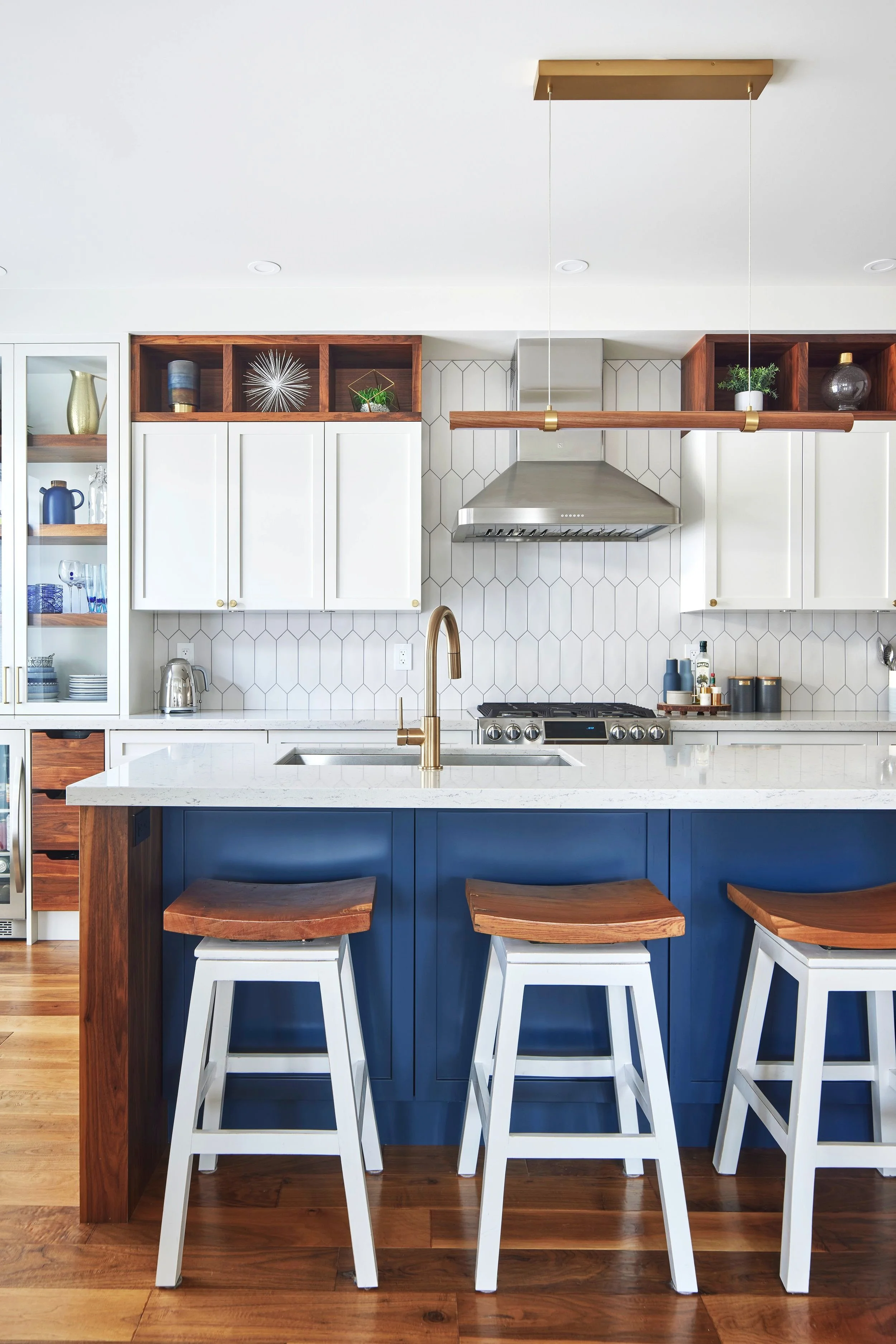 Modern kitchen with dark blue island, white stools with wooden seats, white cabinets, open wooden shelves with decorative items, stainless steel range hood, and hexagonal tile backsplash.