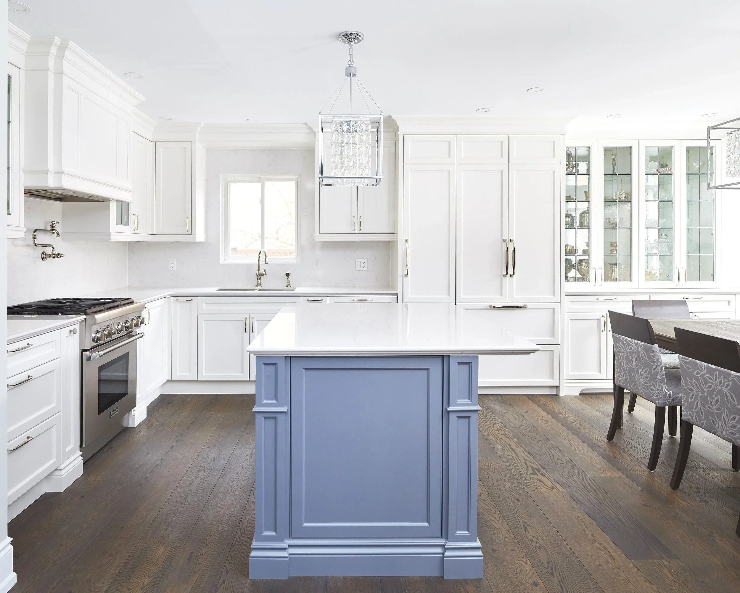 A modern white kitchen with a blue kitchen island, white cabinetry, hardwood flooring, and a crystal pendant light hanging from the ceiling.