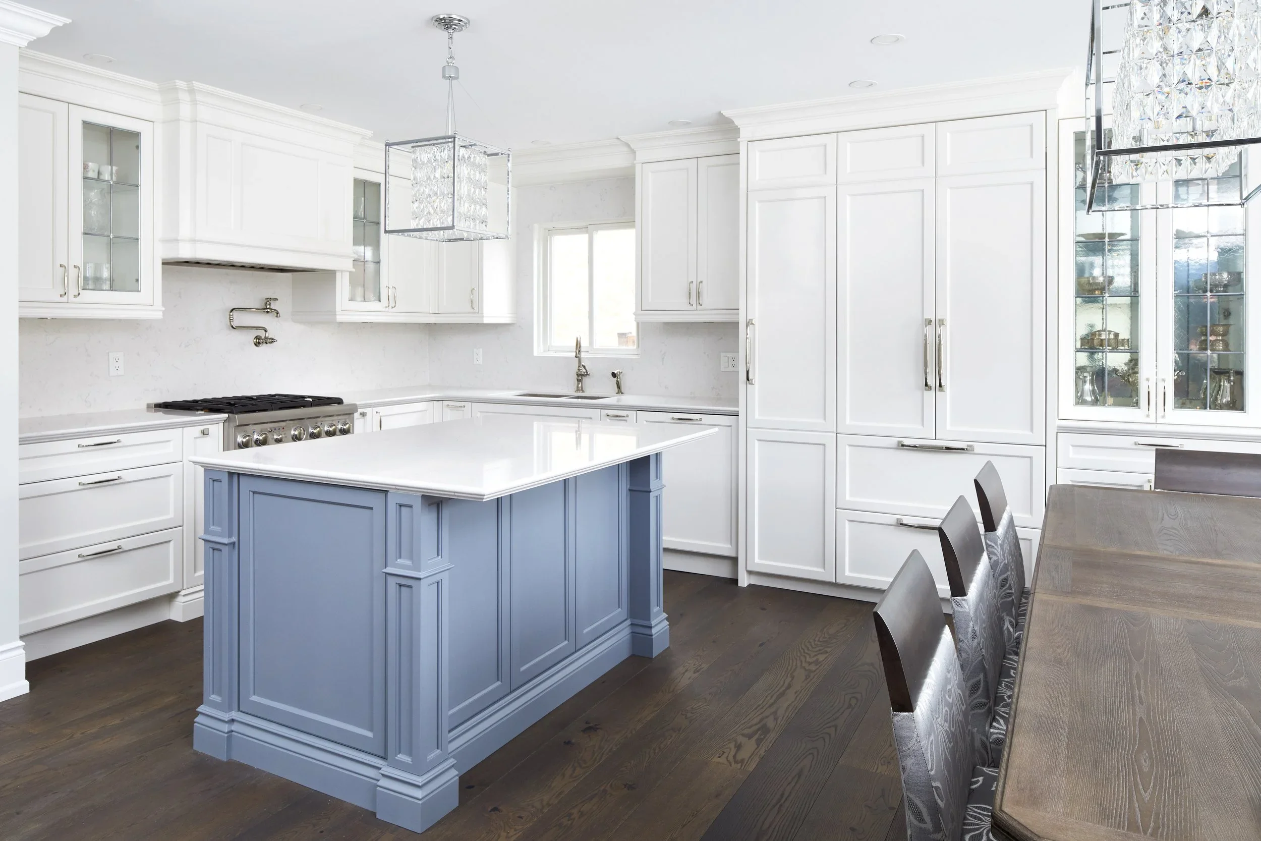 A modern kitchen with white cabinets, a blue kitchen island, and a chandelier. There is a window above the sink and a dining table with chairs.