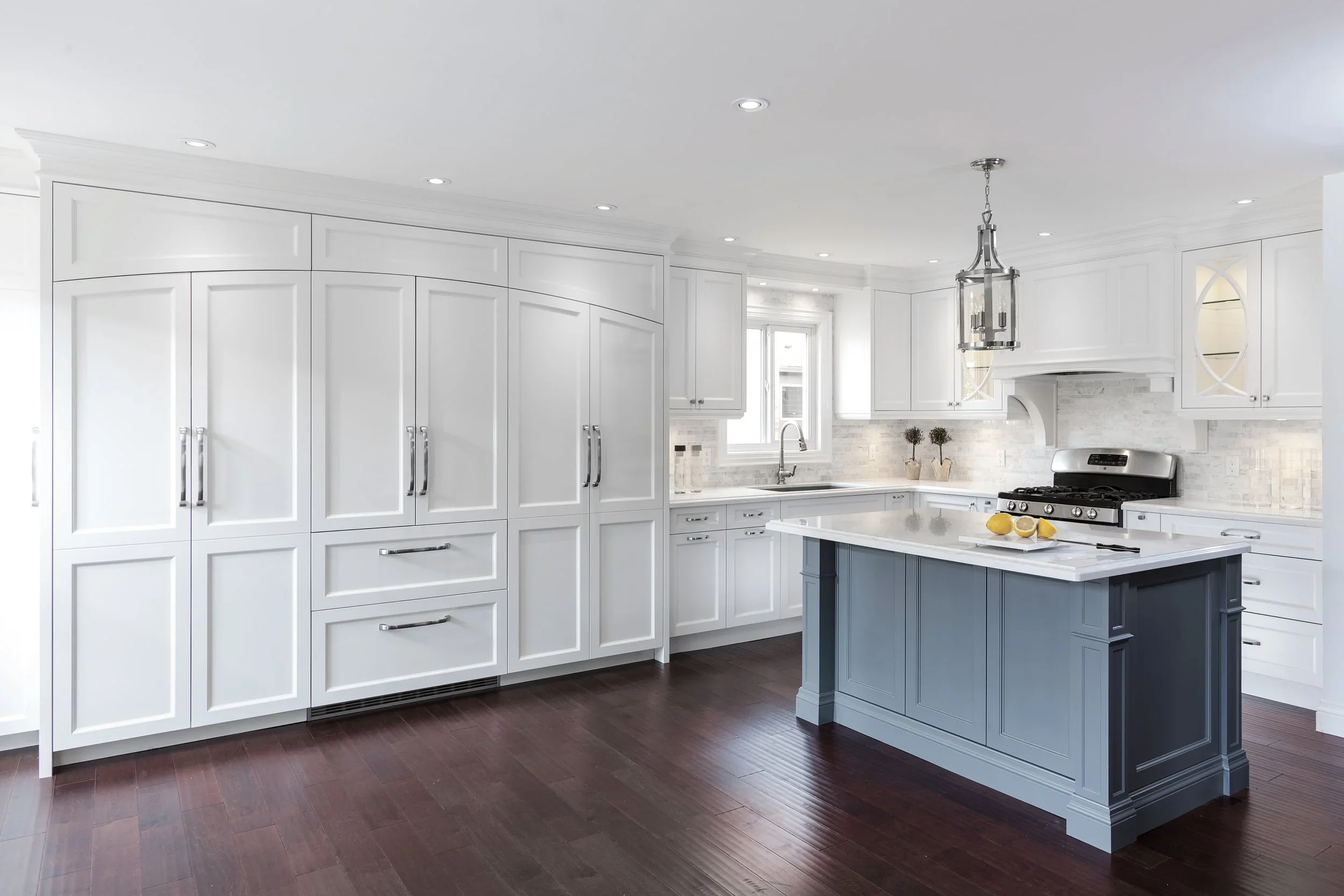 A modern white kitchen with dark hardwood floors, featuring a large island with a light gray base and white countertop, white cabinets, and a stainless steel stove. The space is well-lit with recessed ceiling lights and a hanging light fixture.