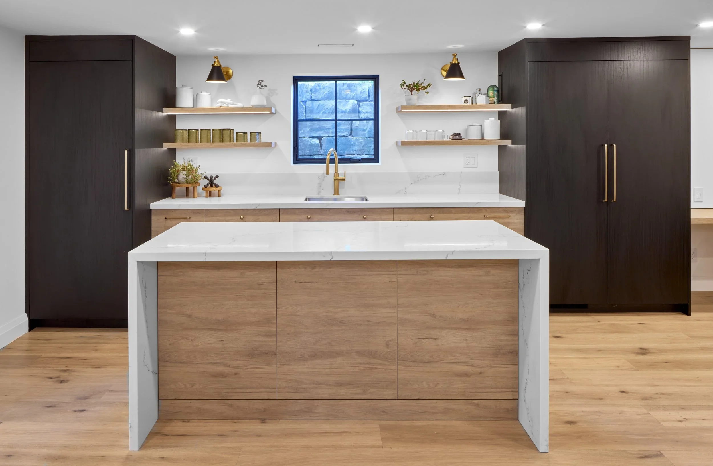 Modern kitchen with dark cabinets, light wood accents, white marble countertops, and a large island. Features small decorative plants, open shelving with jars and plants, and a window with a stone wall outside.