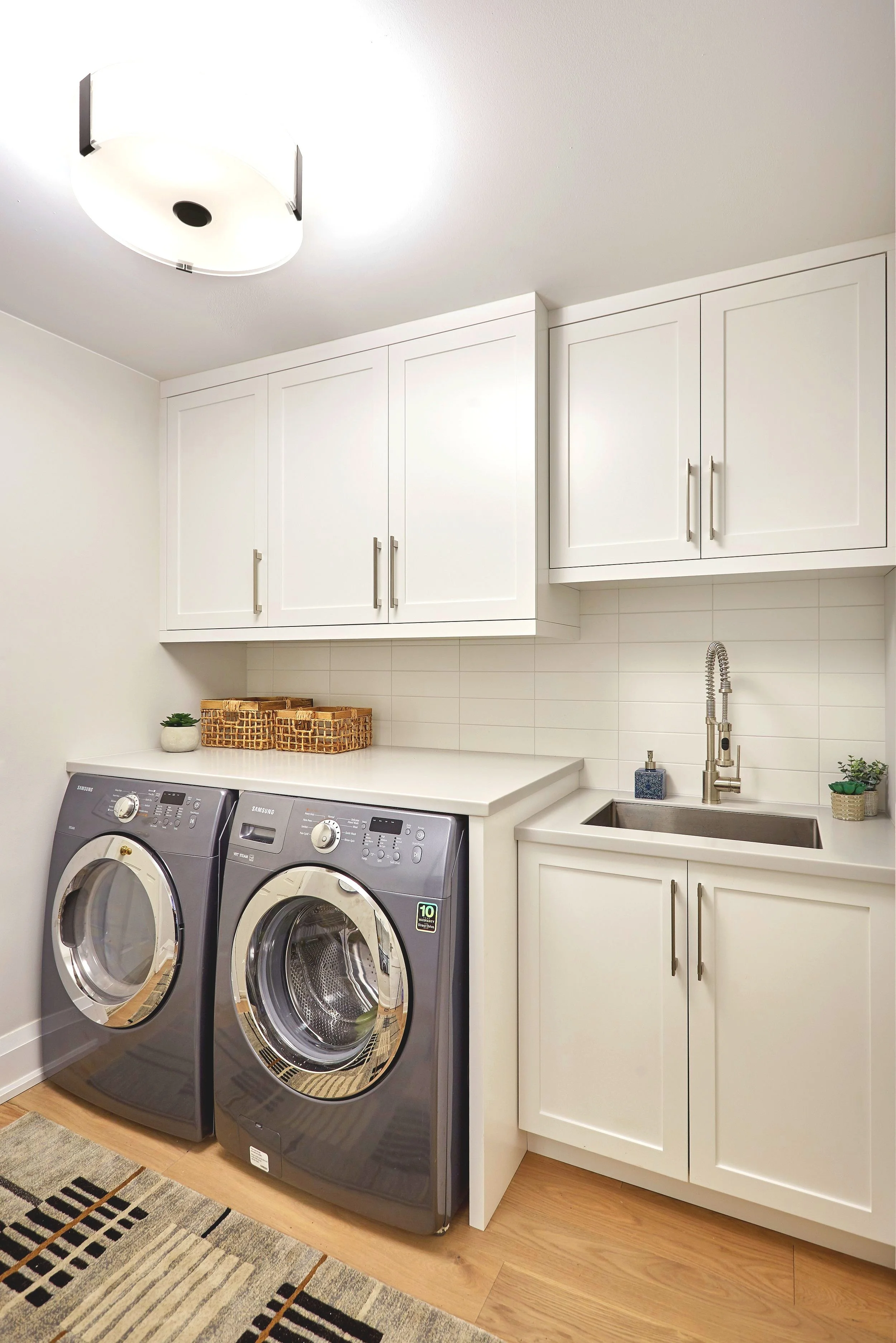 A laundry room with a white countertop, white cabinets, a sink with a faucet, and a pair of front-loading washing machines. Decorative baskets and small plants sit on the countertop. A ceiling light fixture illuminates the room, and a patterned rug is on the floor.