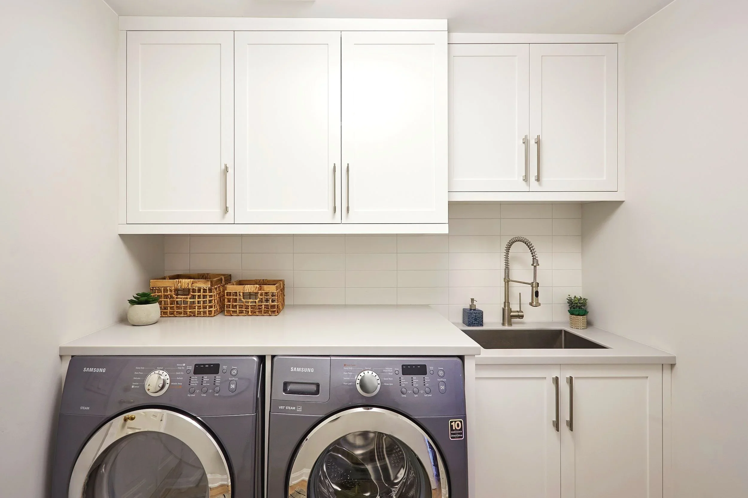 A laundry room with white cabinets above a white countertop, two woven baskets, small green plants, soap dispenser, and a stainless steel sink, with two Samsung front-loading washing machines below.