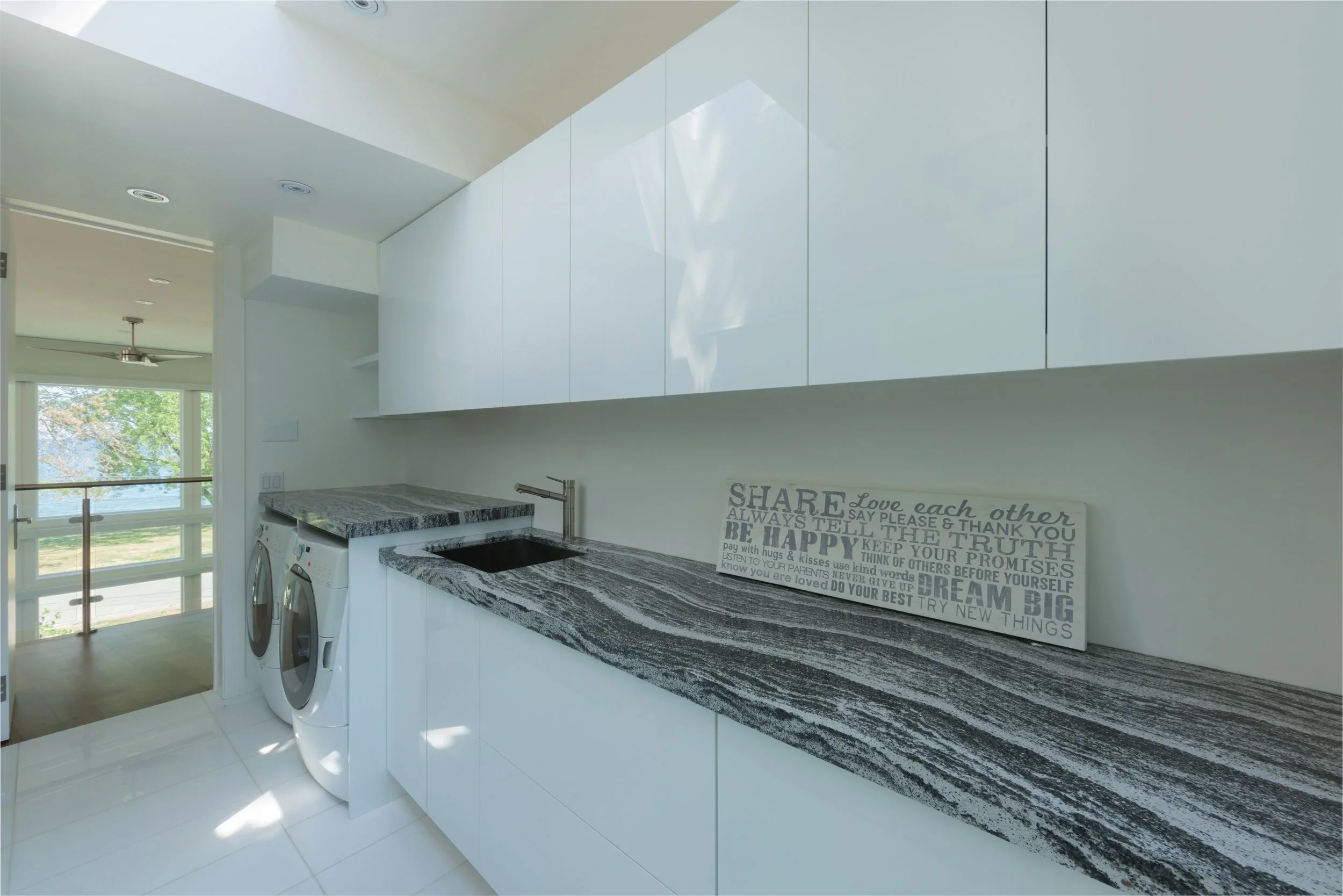 Modern laundry room with white cabinets, granite countertop, and front-loading washer and dryer, with a decorative inspirational quote on the wall, a window with a view of trees, and a ceiling fan.