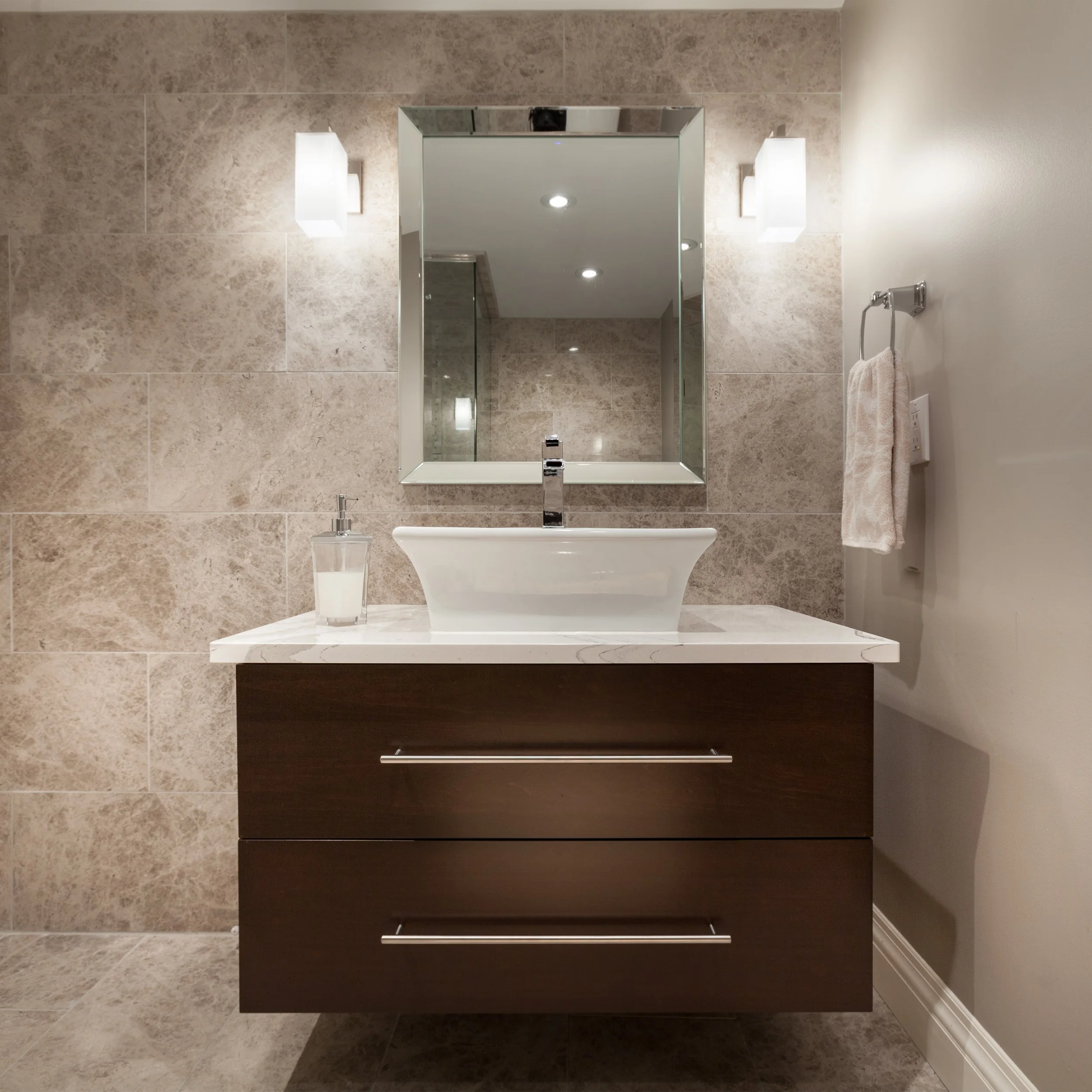 Bathroom mirror with wall lights, sink with a clear soap dispenser, beige tiled wall, and a towel hanging on a holder.