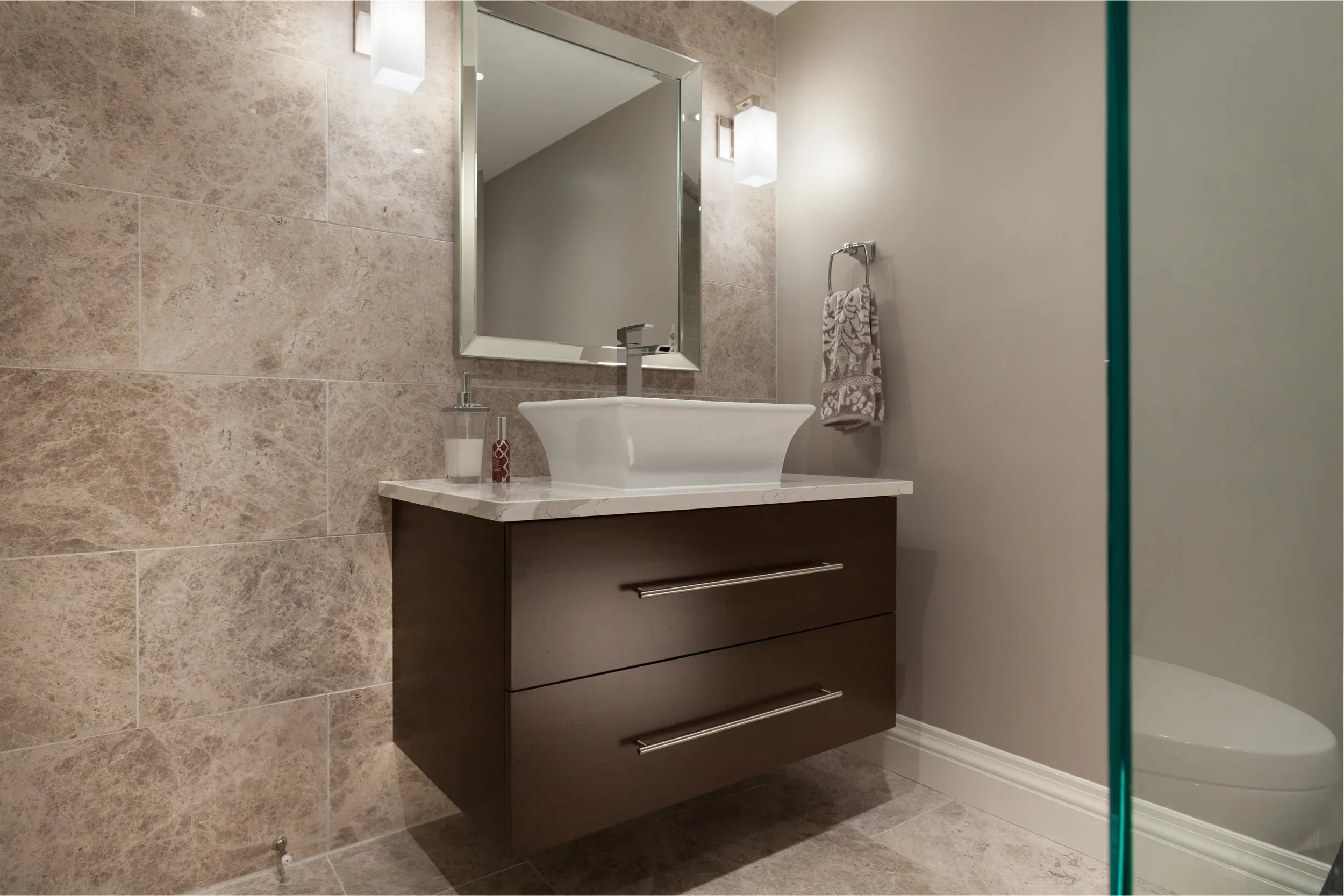 Bathroom with beige tiled walls, a white sink, and a dark brown cabinet with three drawers. There is a large rectangular mirror above the sink, a soap dispenser, and a towel hanging on a circular towel ring. Part of a shower with a glass door is visible on the right side.