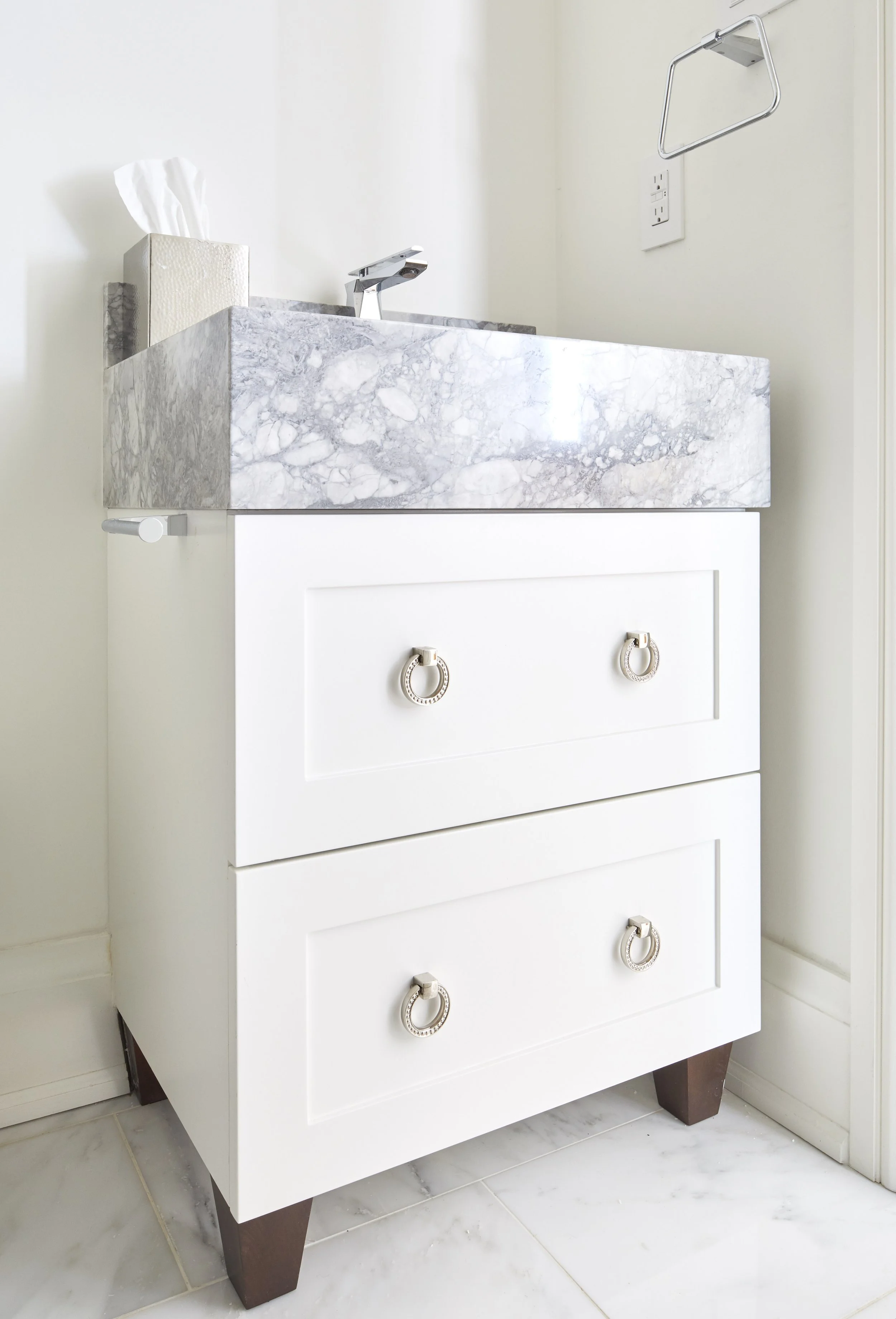 A white bathroom vanity with a marble countertop, a faucet, tissue box, towel holder, and decorative drawer handles, set against a light-colored wall and tiled floor.
