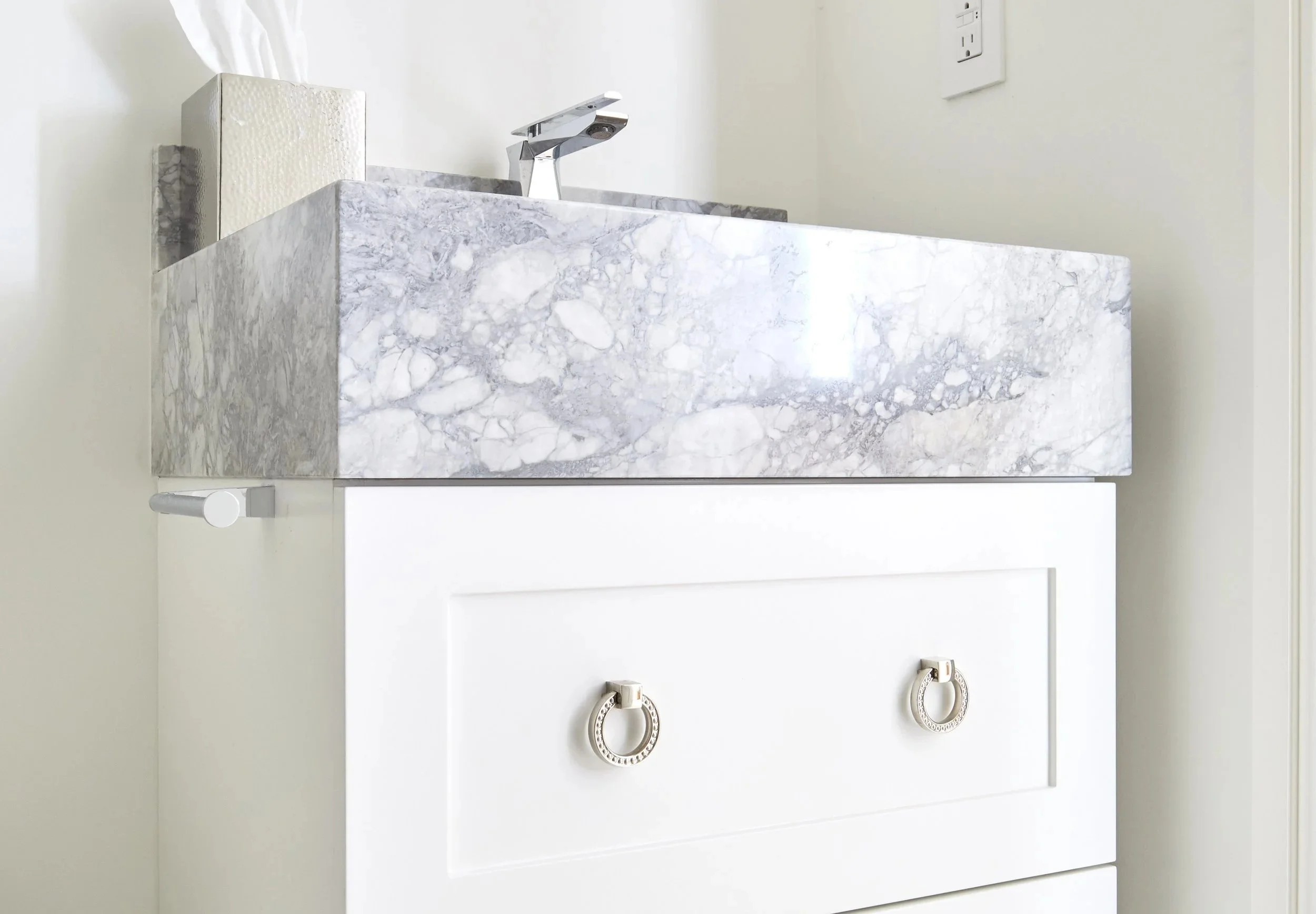 Close-up of a marble countertop with a modern chrome faucet and a tissue box, above a white cabinet with two decorative round handles.