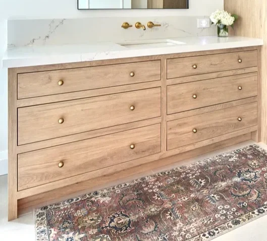 Bathroom vanity with six drawers, a marble countertop, a gold faucet, a mirror, a vase with white flowers, and a patterned rug on the floor.