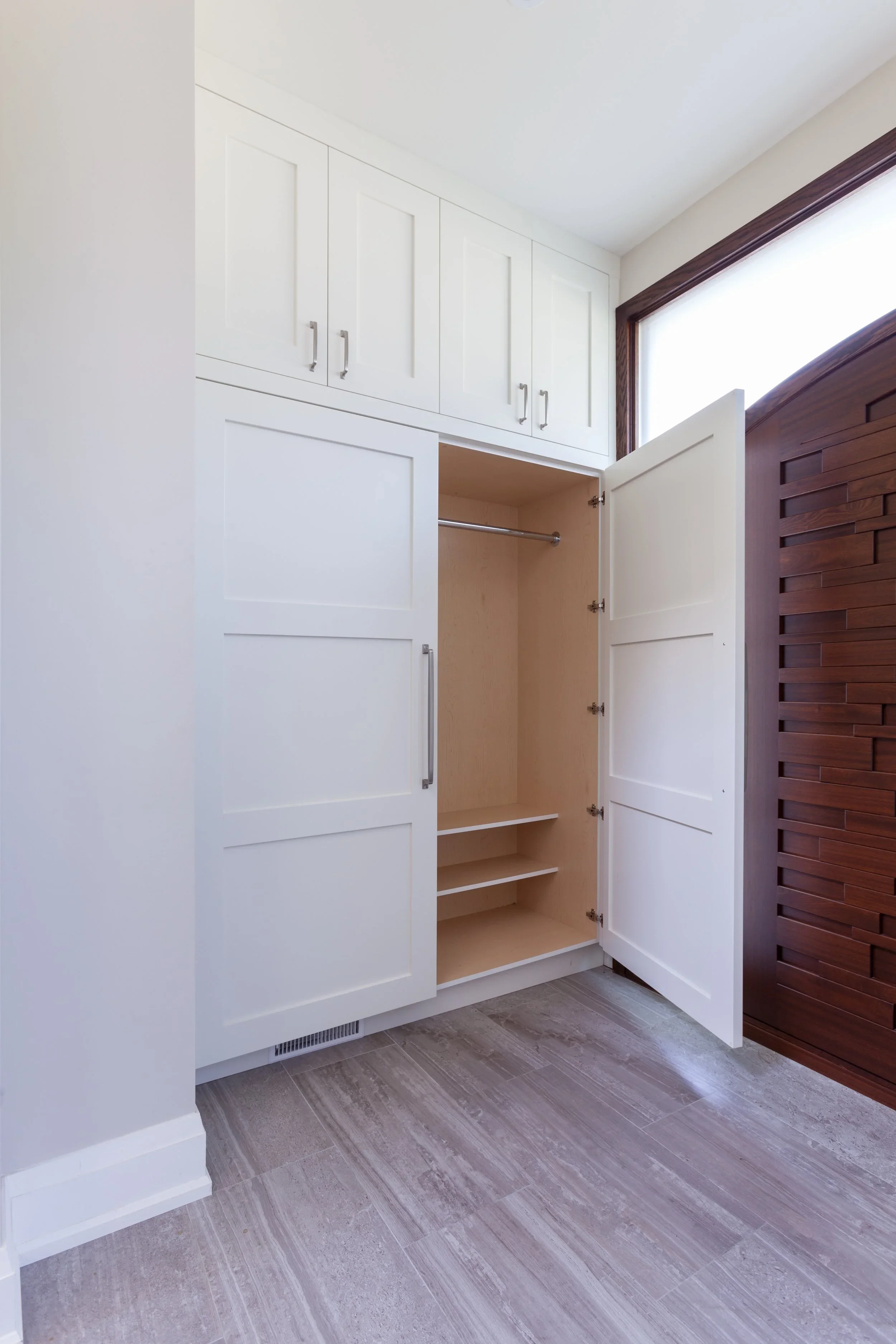 Open white built-in closet with upper and lower cabinets, and empty interior with a hanging rod. Brown wood trim around a large window with frosted glass on the right, and light gray wood-look tile flooring.