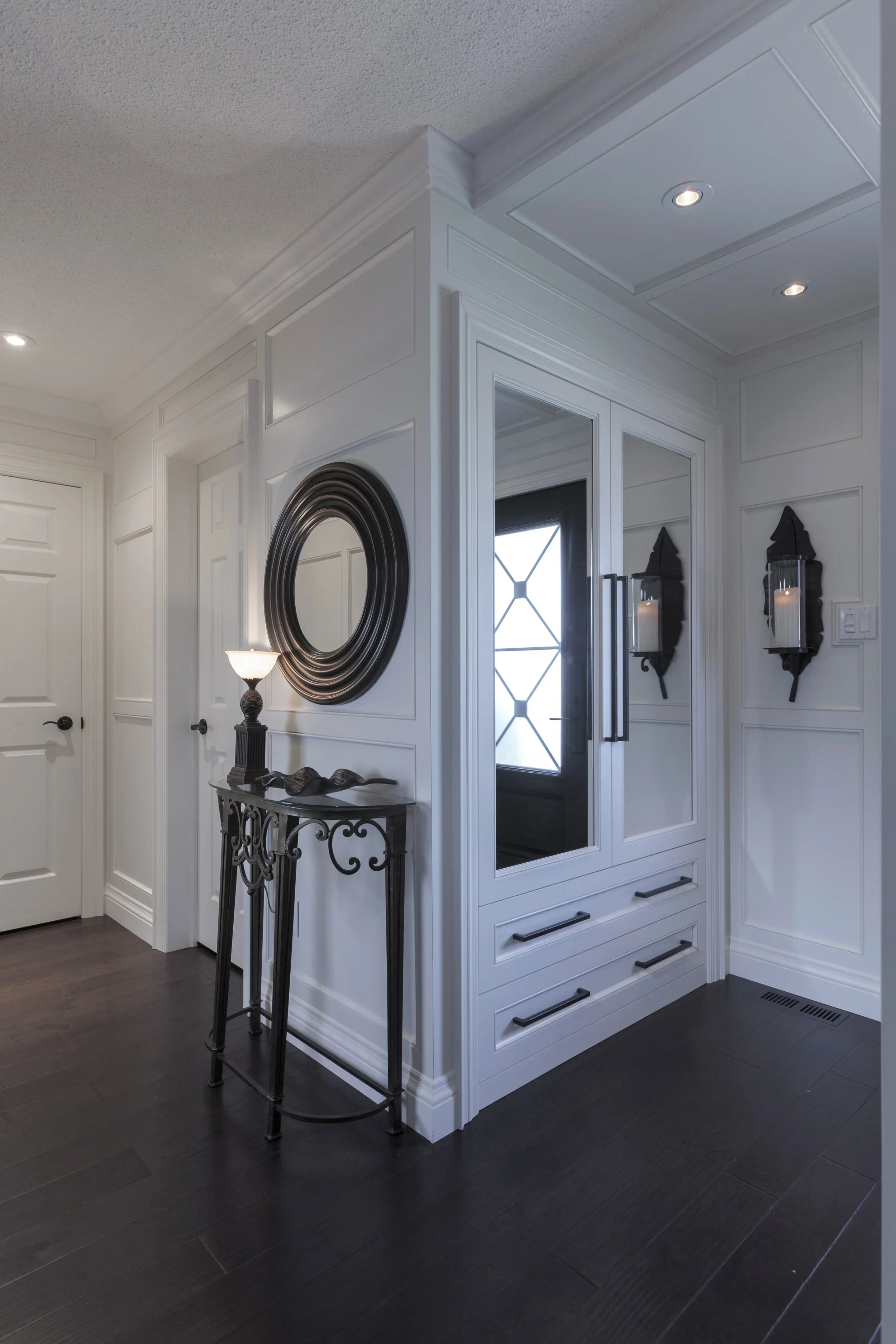 Entryway with white paneled walls, black door with glass insert, black wall sconces with candle candles, decorative black metal table, round black mirror, and dark wood flooring.