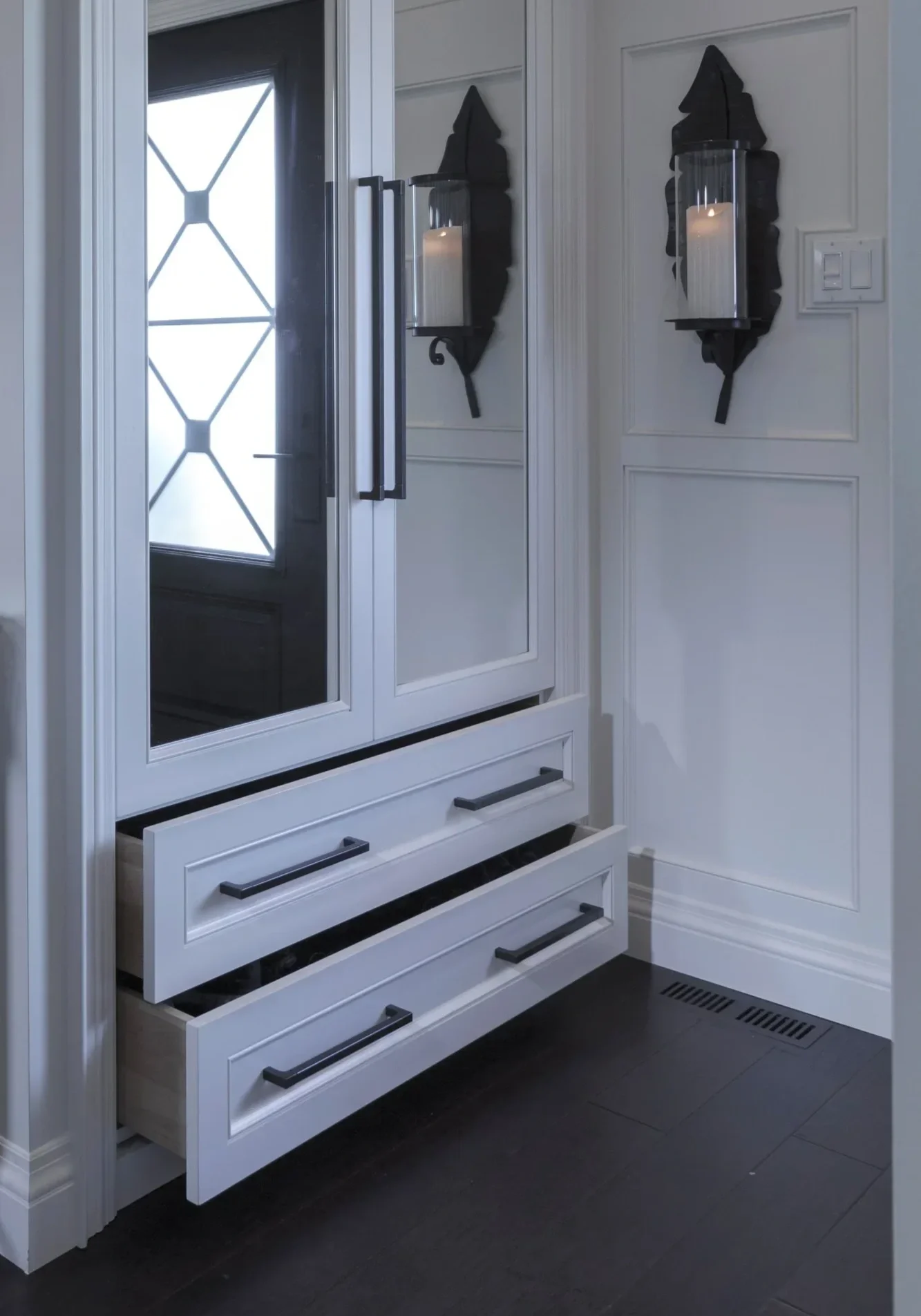A white cabinet with black handles and open drawers, situated in a corner of a room. On top of the cabinet, there is a large mirror and two wall-mounted candle holders with glass covers, each containing a lit candle. The room has white walls with decorative molding, a door with a glass panel, and a black floor vent.