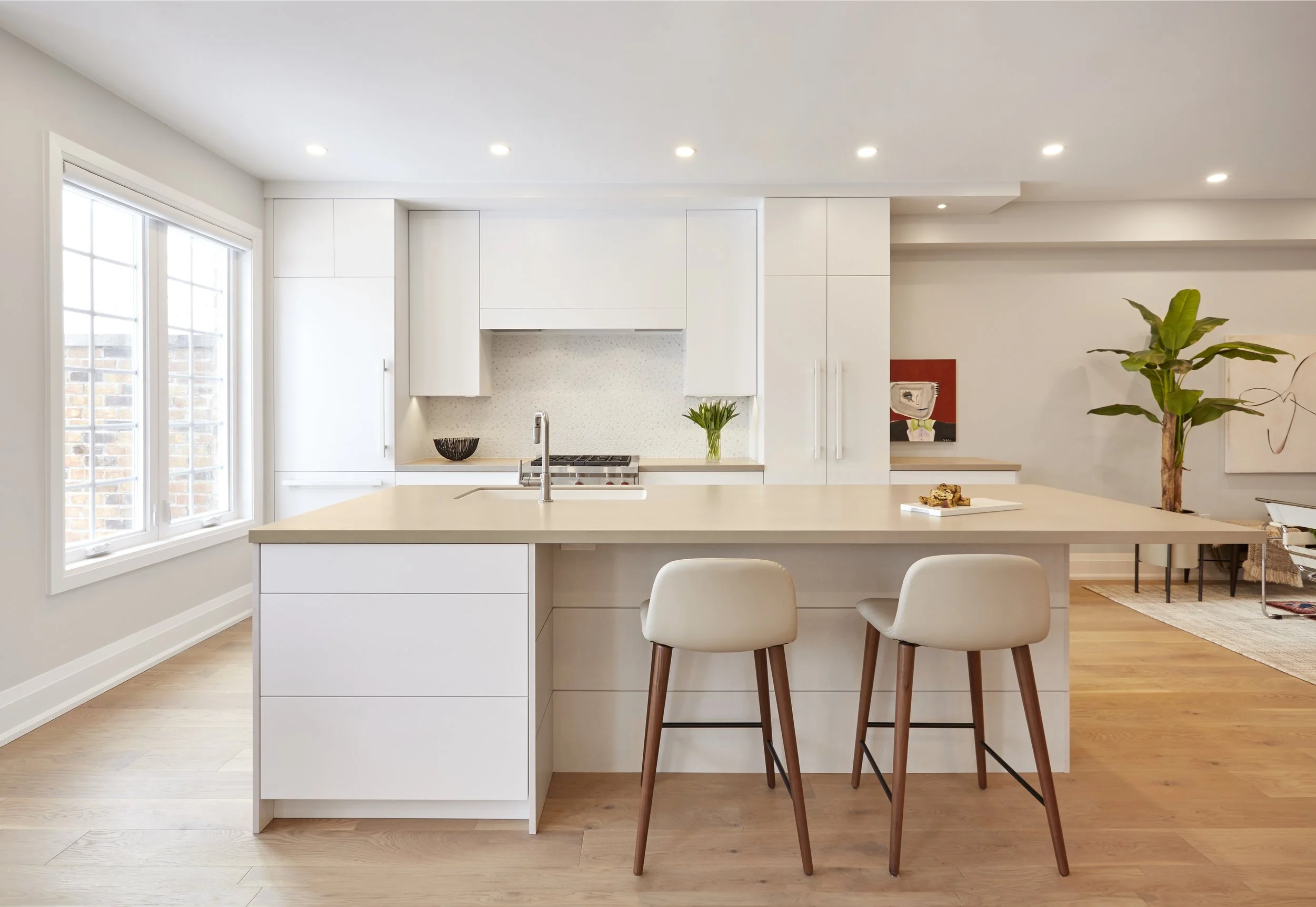 Modern kitchen with white cabinets, a beige countertop, two bar stools, a small potted plant, and natural light from large windows.