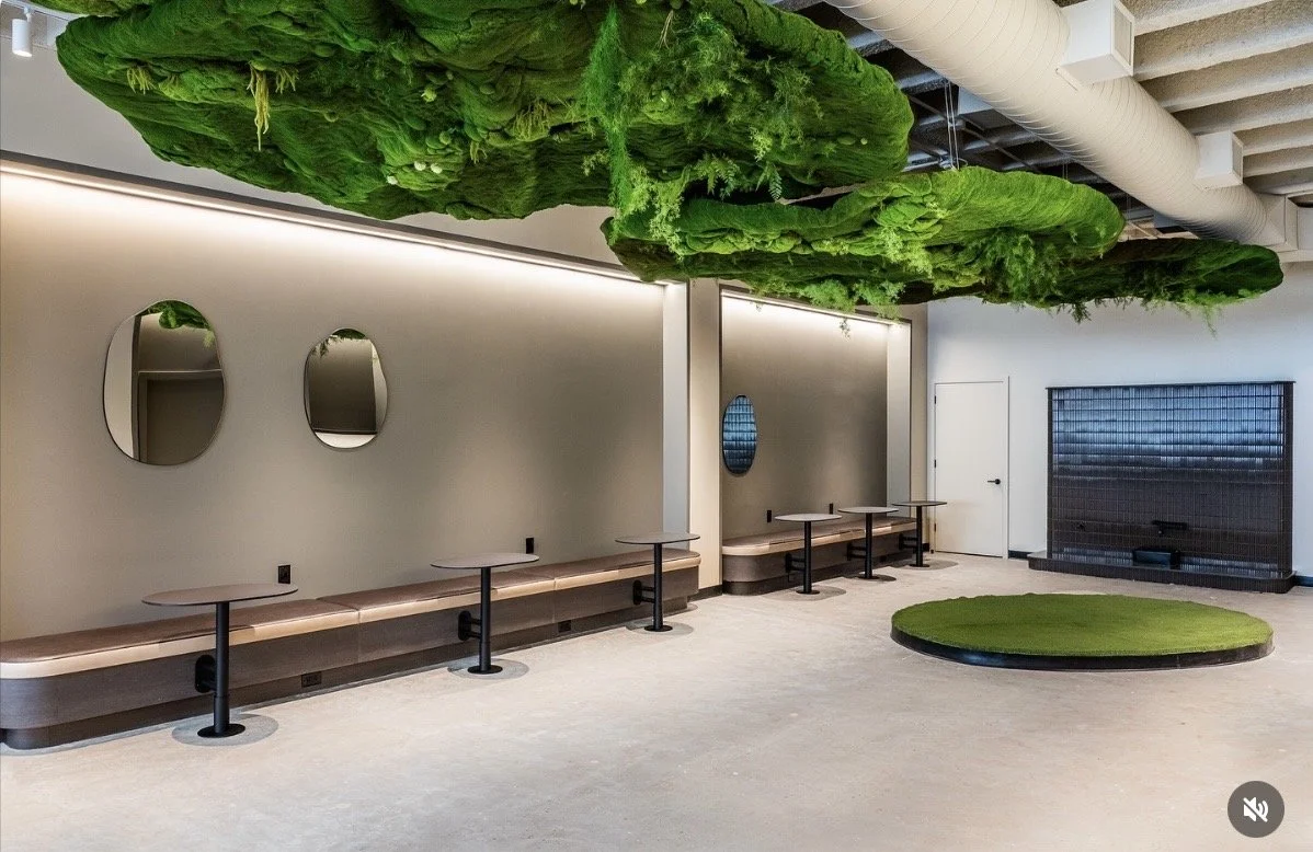 Modern interior space with circular green moss ceiling installations and a moss-covered circular platform on the floor. There are four small tables with stools along the wall, which has three round mirrors. The room has a minimalist design with neutr
