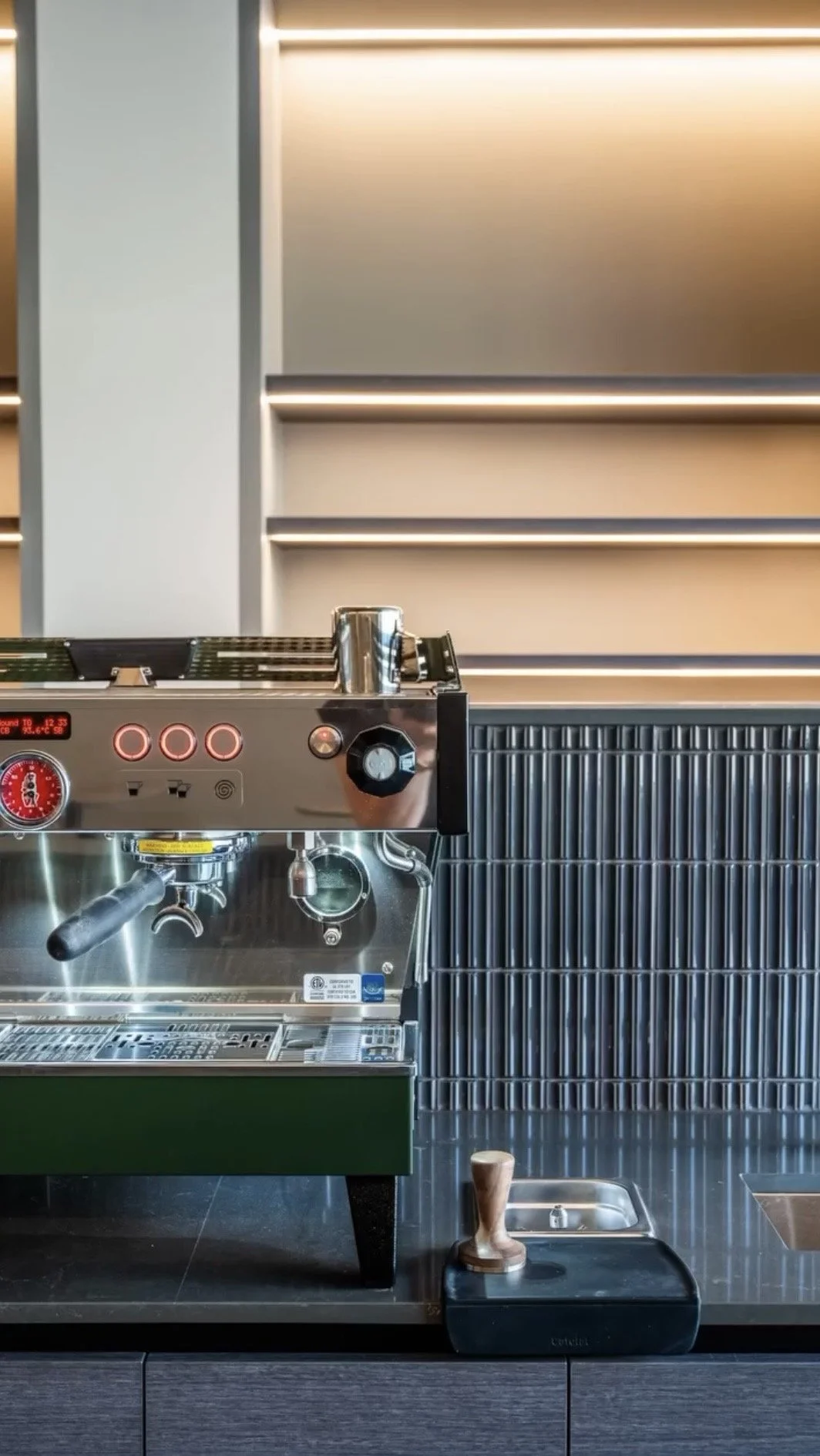 A commercial espresso machine on a black countertop, with a milk pitcher on top, a small sink, and a tamper nearby, against a modern, minimalist background with horizontal lighting.