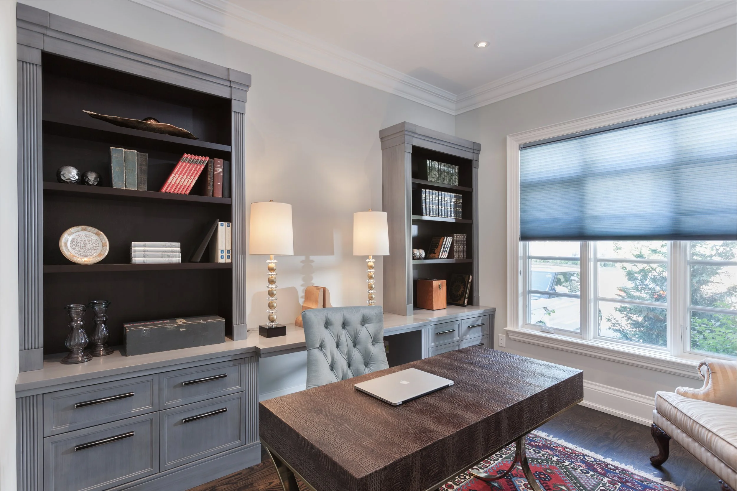 A dining room with blue-gray built-in shelves, a large window with a blue roman shade, and a wooden table with a laptop and a patterned runner. There are two table lamps and a chair.